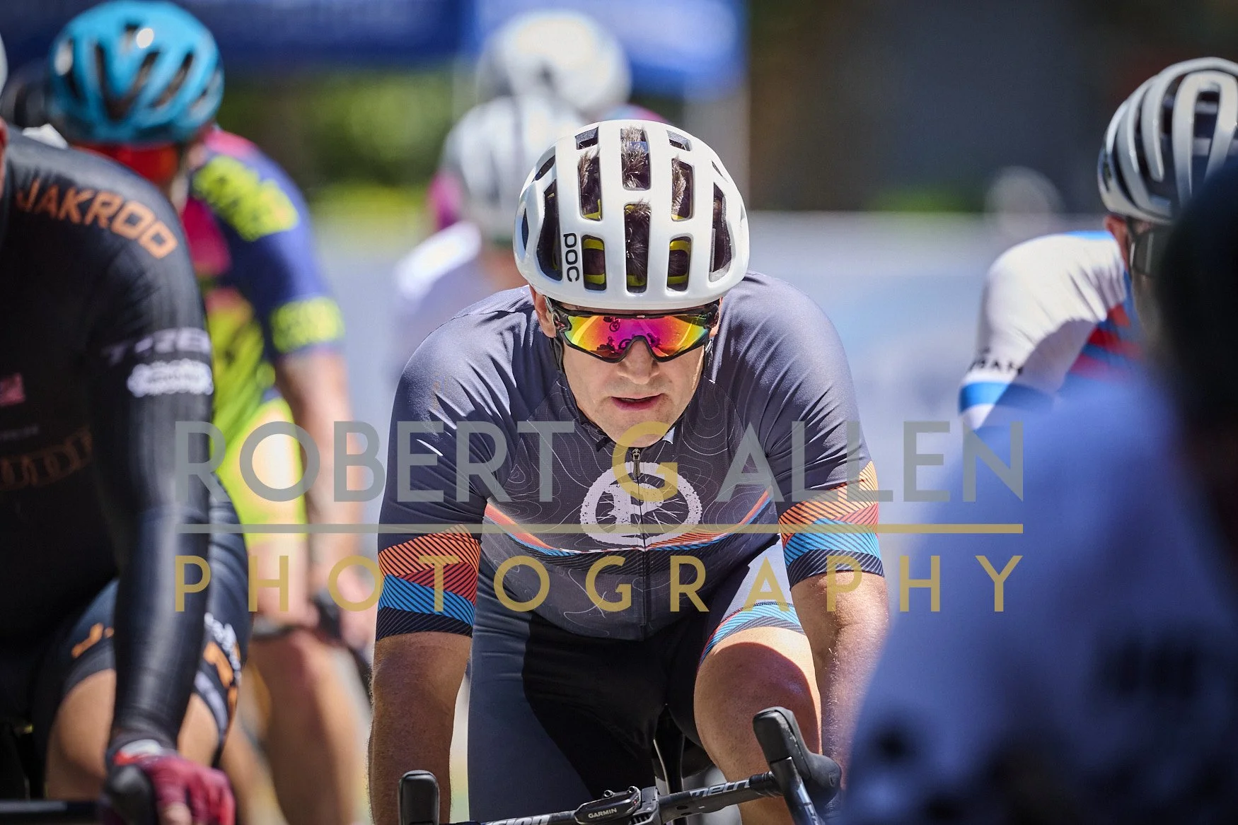 Close-up of a male cyclist wearing a white helmet with black ventilation slots, reflective sunglasses, a dark cycling jersey with colorful stripes on the arms, and gripping the handlebars of his bike during a race. Other cyclists in colorful jerseys 