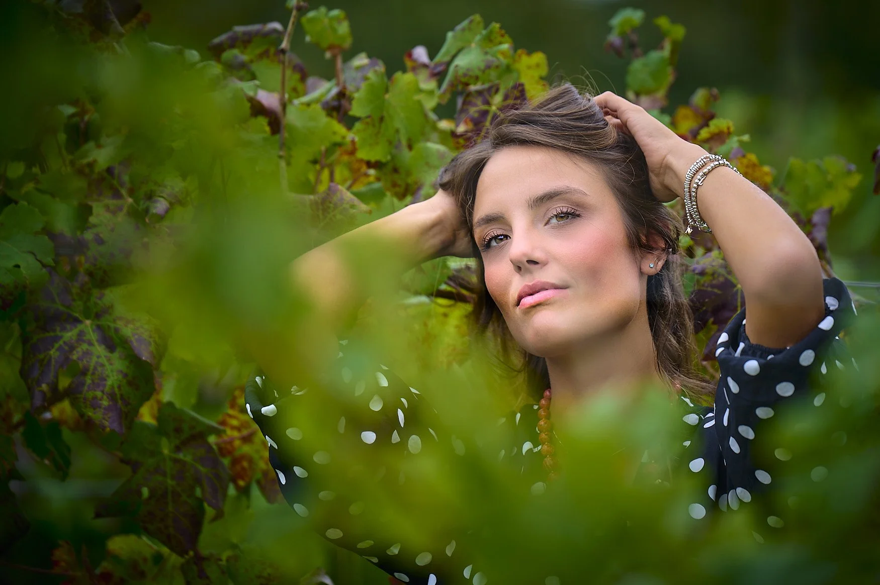 A young woman with blonde and brown hair, wearing a polka dot dress, jewelry, and a red necklace, smiling with her hands behind her head among green grapevine leaves.