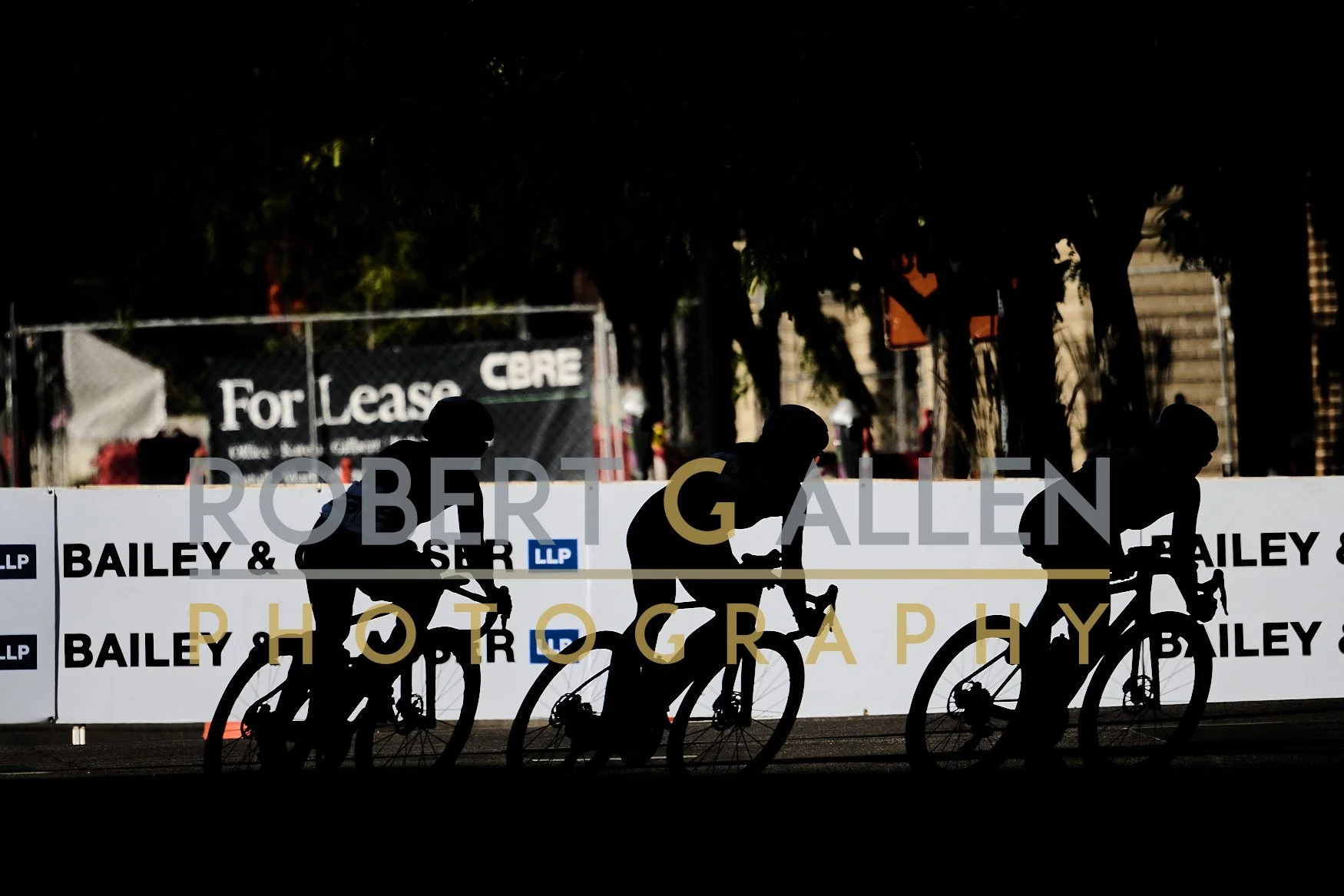Silhouettes of four cyclists riding on a track with a white barrier displaying text and logos, background includes trees and a fence with a 'For Lease' sign.