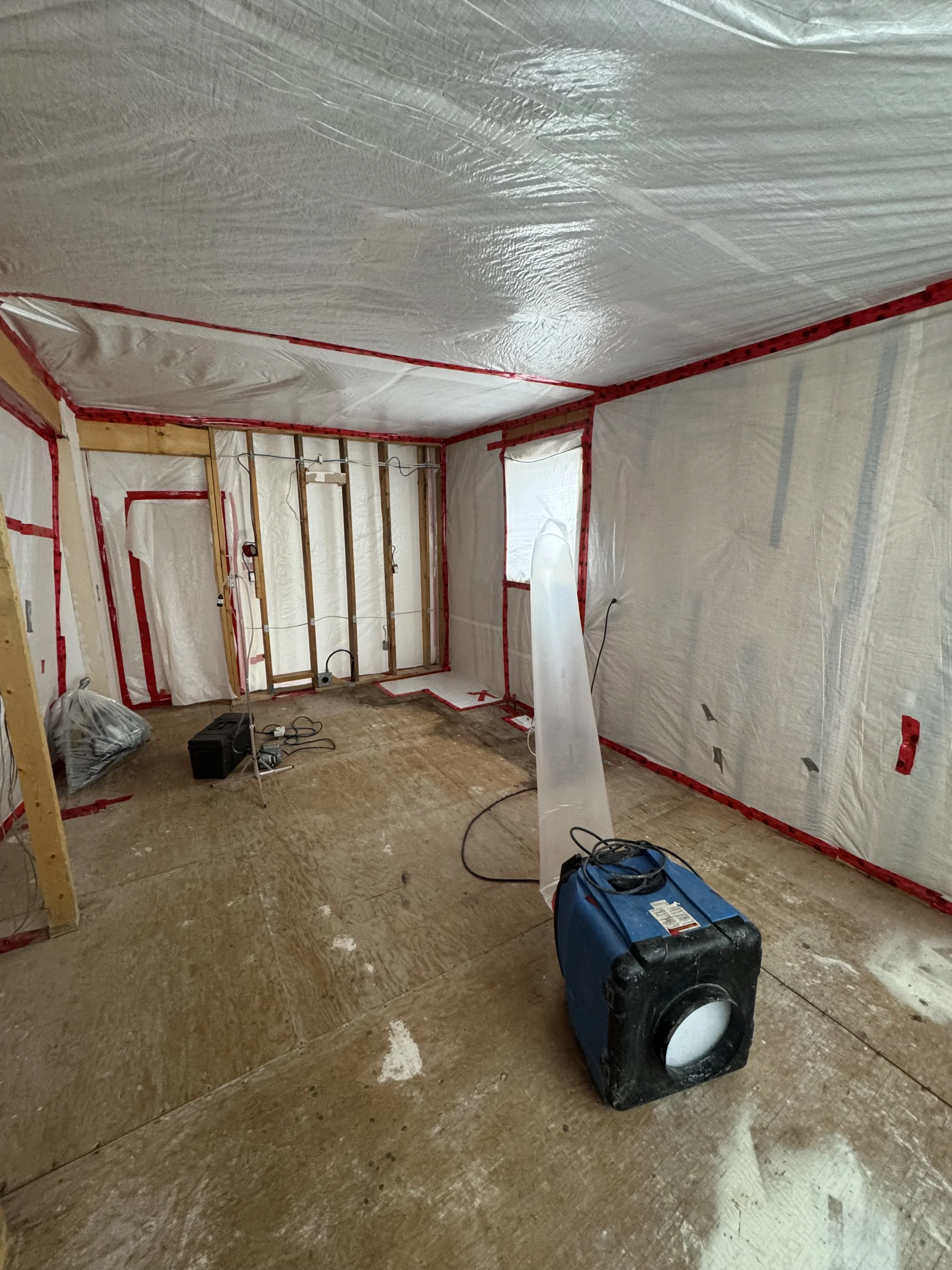 Interior of a room under renovation, with exposed wall studs, plastic sheeting on the walls and ceiling, construction tools, and a portable speaker on the floor.