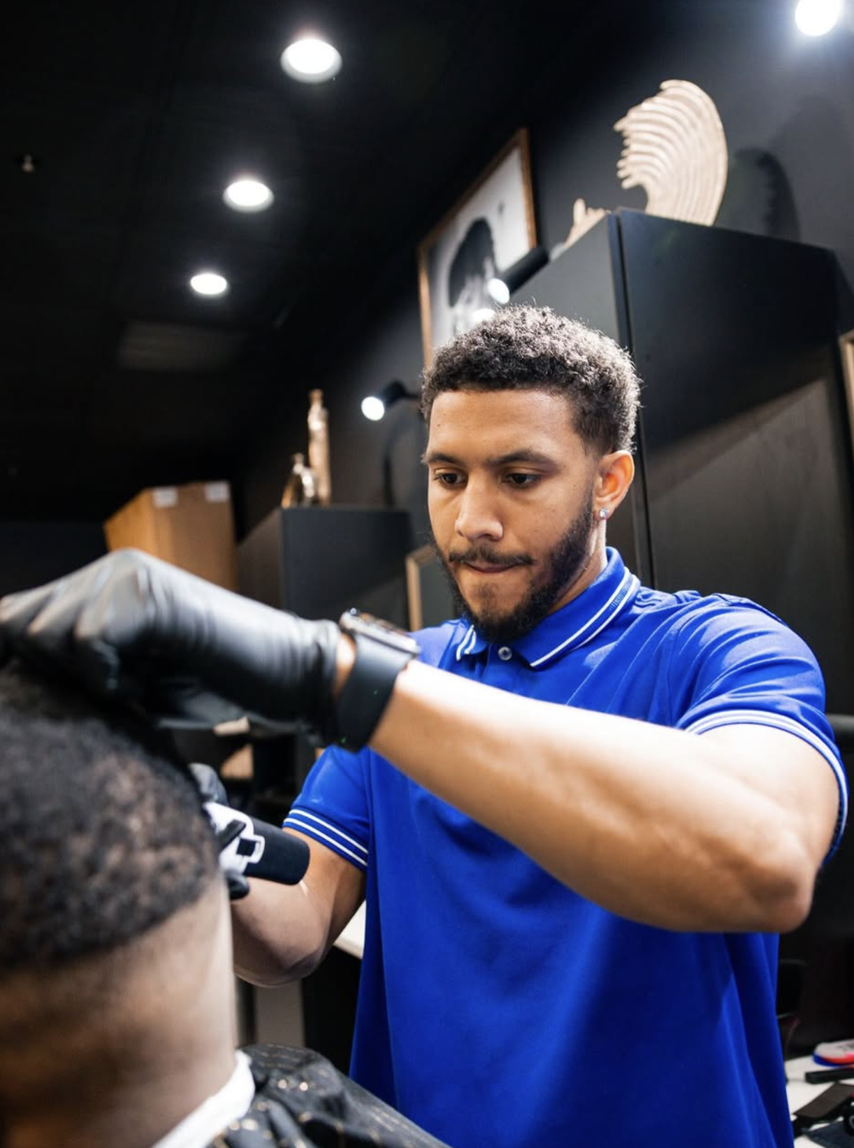 Barber trimming a customer's hair in a barbershop with black walls and framed pictures.