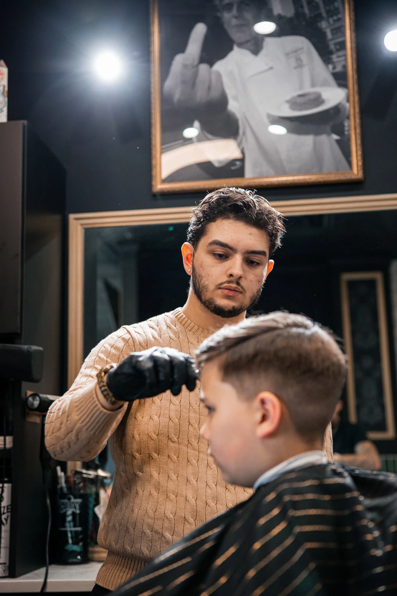 A barber cutting a man's hair in a barbershop, with black walls, gold-framed mirrors, and barber supplies visible in the background.