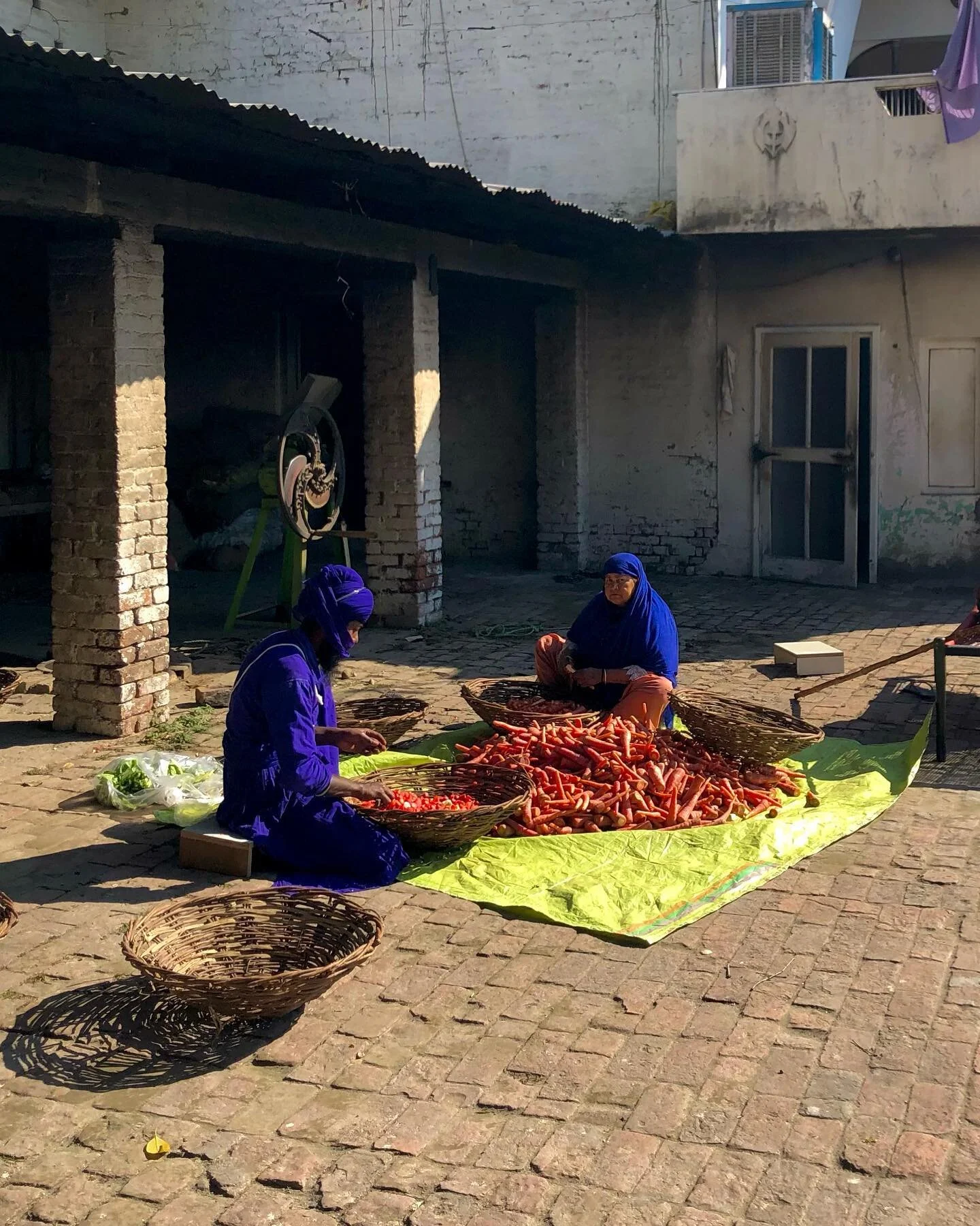 outside the verundah of ਸ਼ਹੀਦੀ ਬਾਗ ਸਰਾਂ ('martyr's garden' serai) a pair of persons do the honorable service of washing and preparing carrots &mdash; popularly winter's harvest in the Anandpur Region &mdash; lunger for visitors and residents of the b