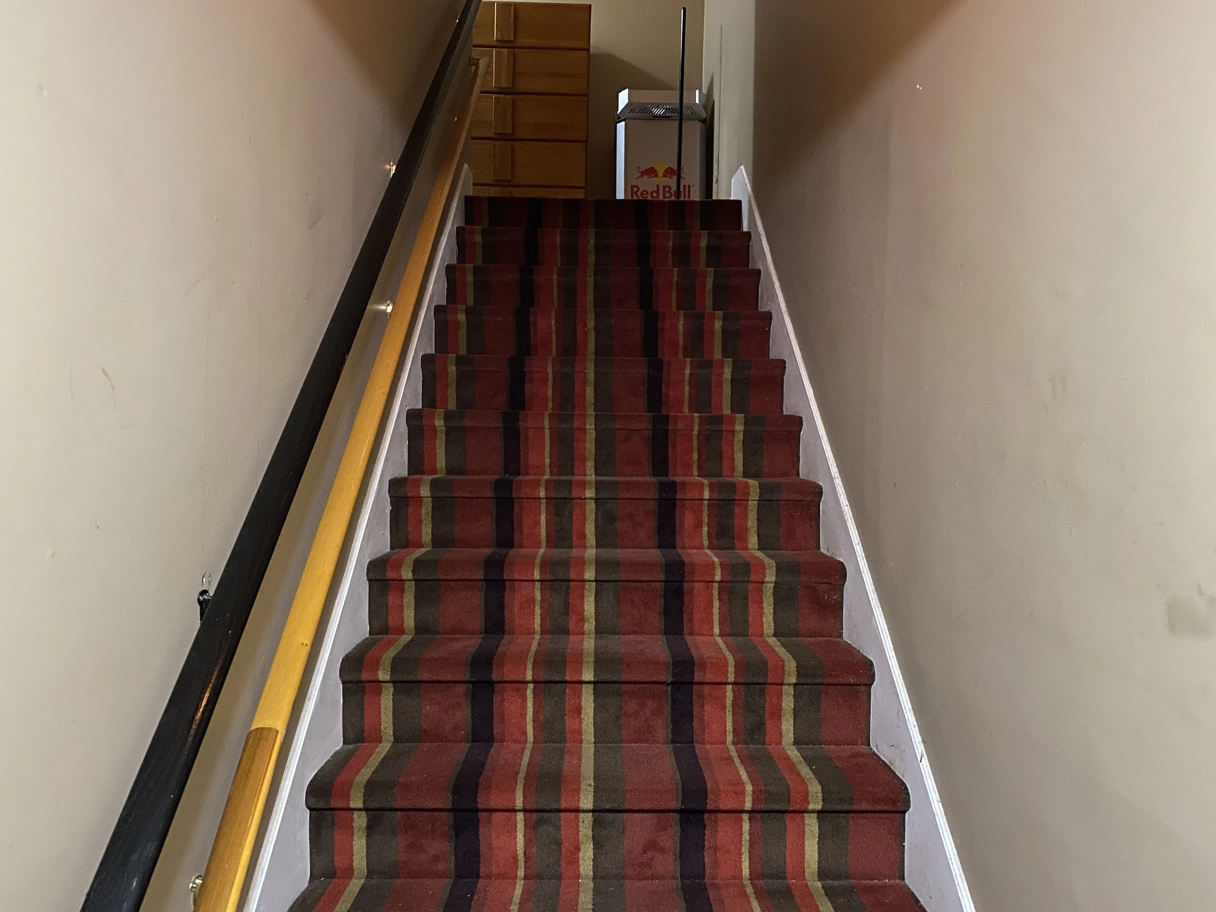 Interior staircase with a multi-colored striped carpet, a yellow handrail on the left, and a wooden dresser at the top.