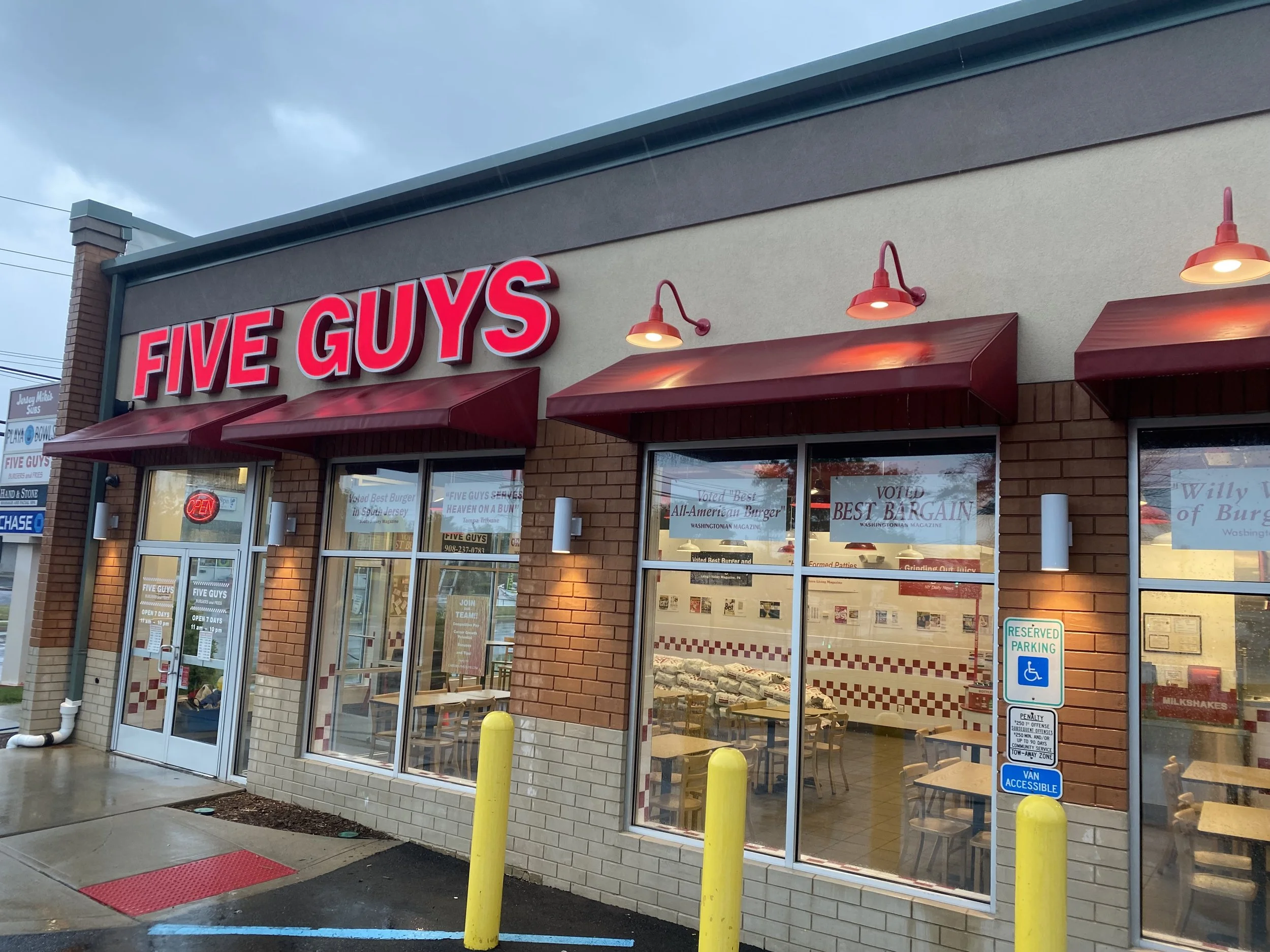 Exterior of a Five Guys burger restaurant with red sign and red awnings, rain puddles on the sidewalk, and parking signs in front.