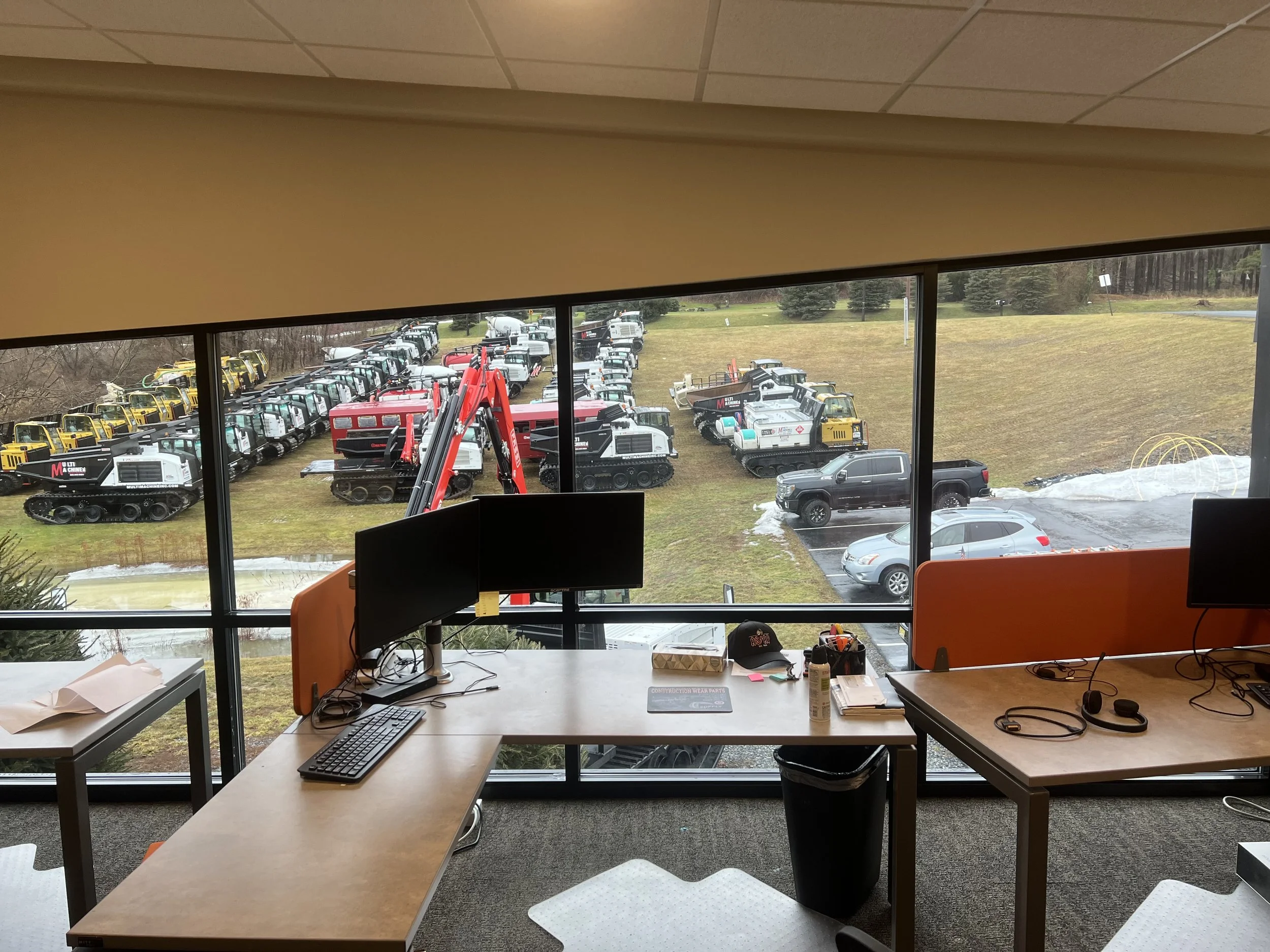 View from inside an office showing a large window overlooking a parking lot and a construction site with small excavators and bulldozers.