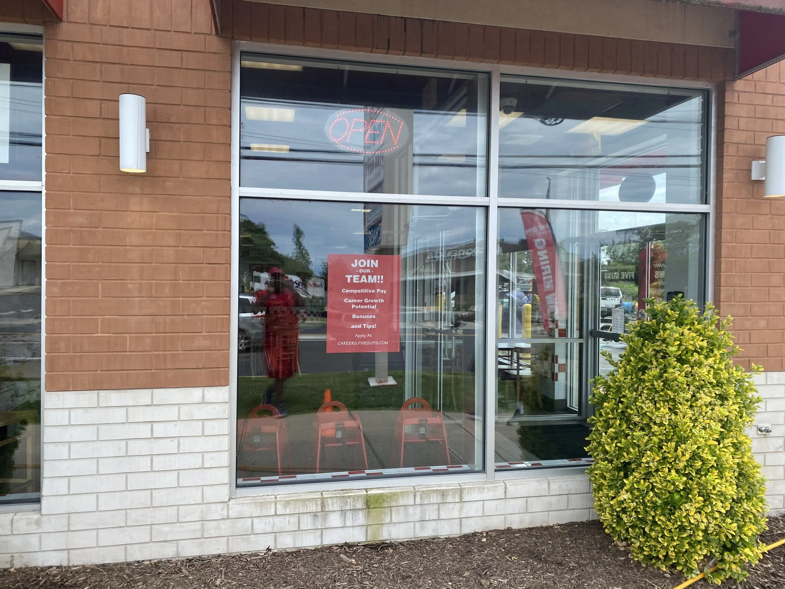 The exterior of a restaurant with a glass storefront, a bright green shrub outside, a red open sign inside, and a red poster promoting job opportunities on the window.