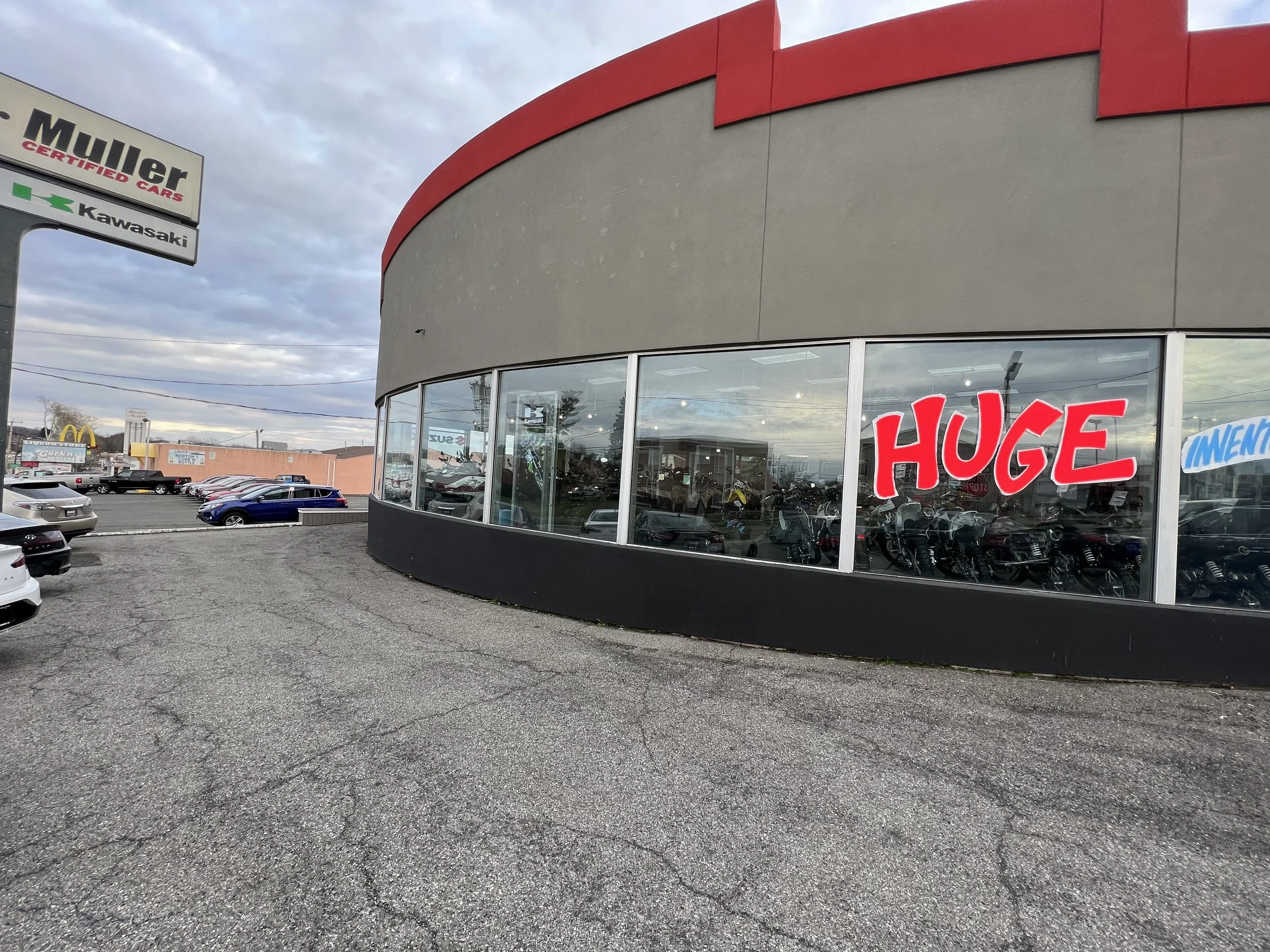 Car dealership with a large red sign on the window saying "HUGE" and a car lot with several vehicles, including a McDonald's sign and a McDonald's restaurant in the background.