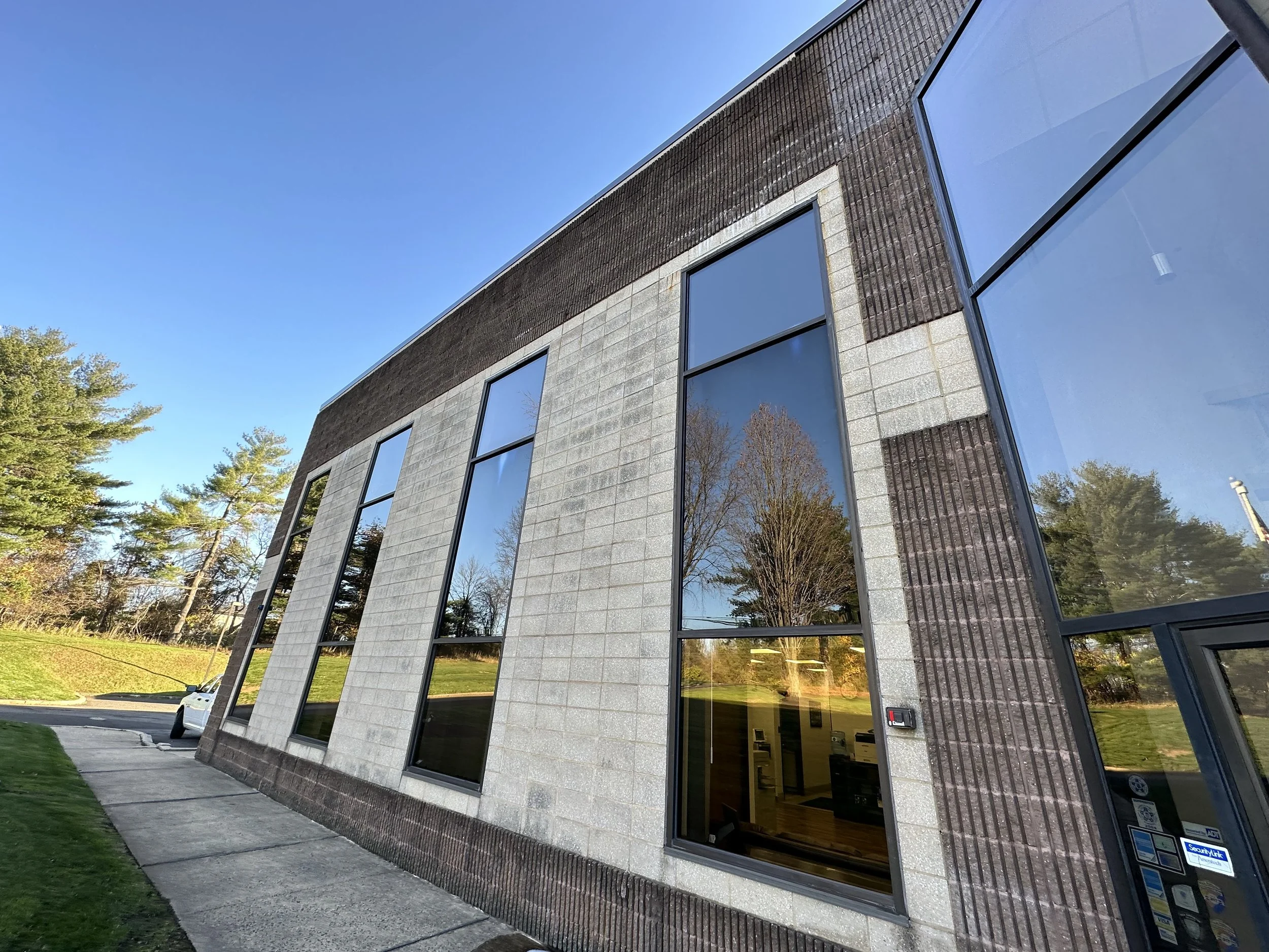 Modern commercial building with large glass windows reflecting trees and a clear blue sky, with a sidewalk and lawn in the foreground.