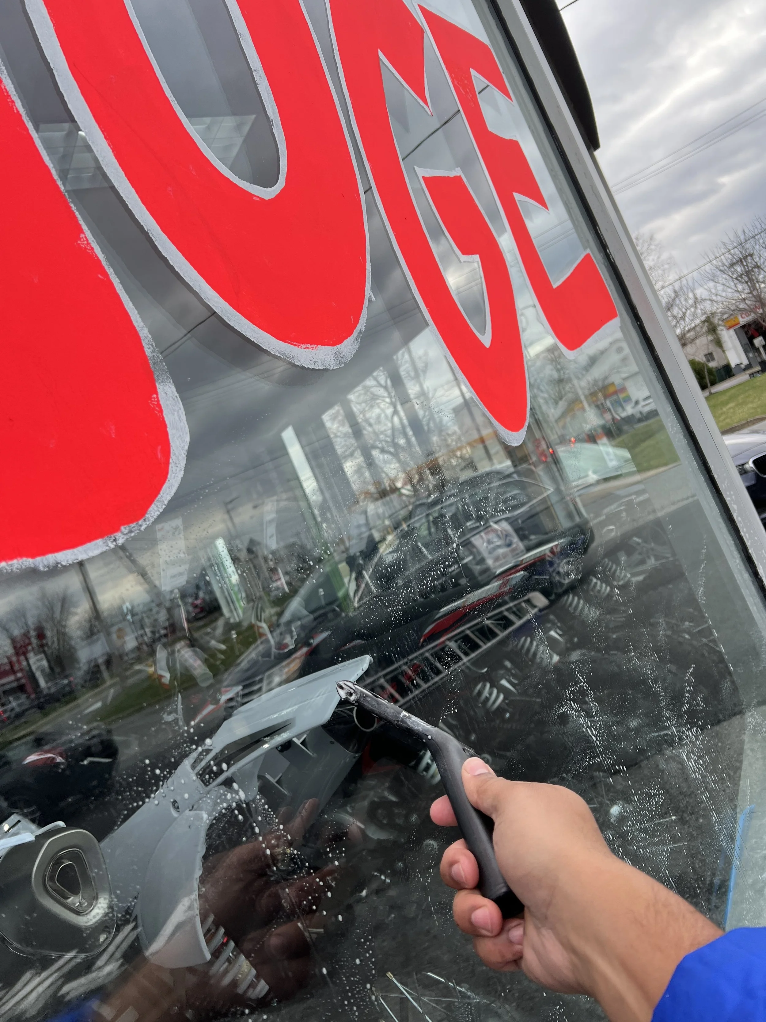 A person cleaning the glass window of a store with a squeegee, with a large red and white 'Open' sign on the window, and cars visible through the window.
