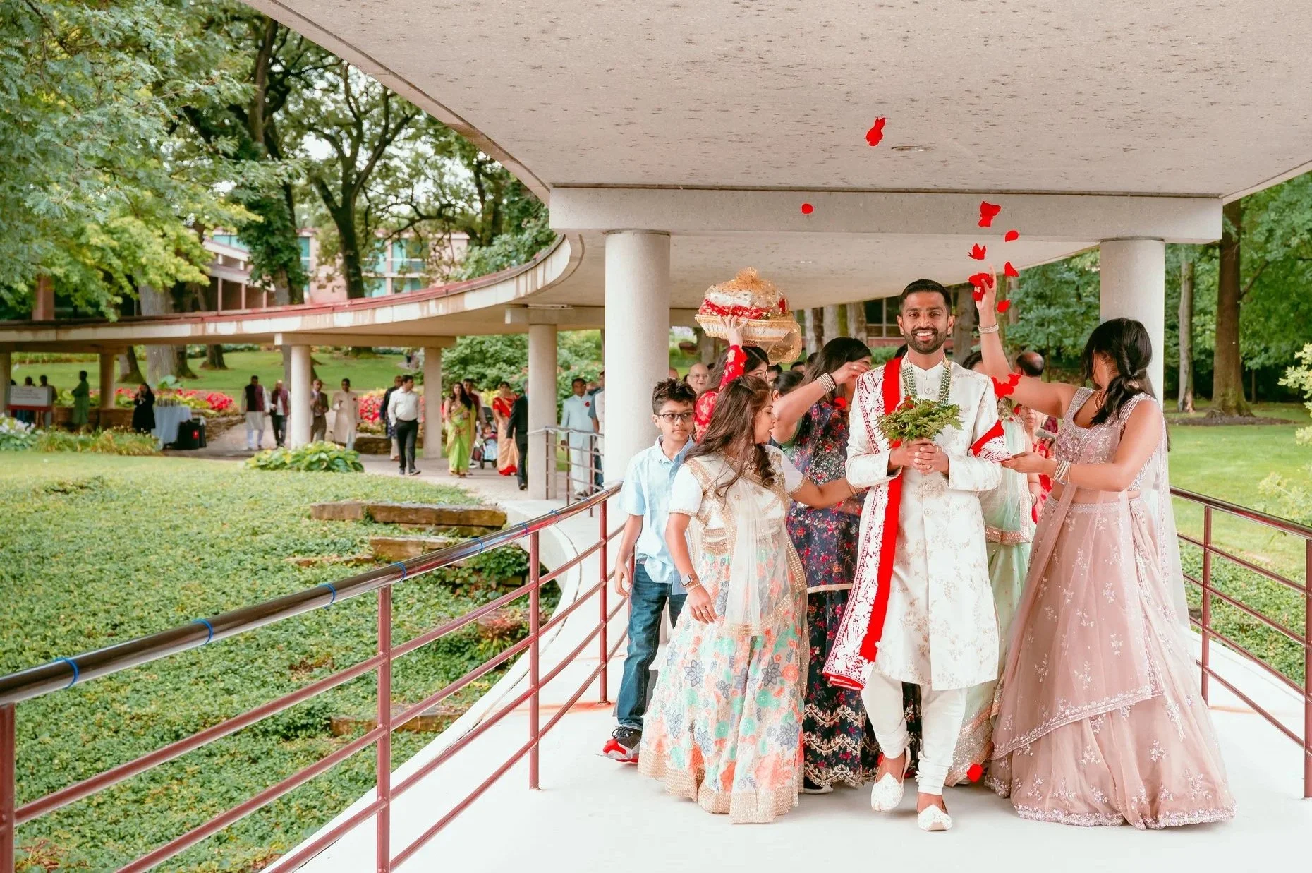 A wedding procession with a groom holding a bouquet, surrounded by family and friends, throwing flower petals in a park.