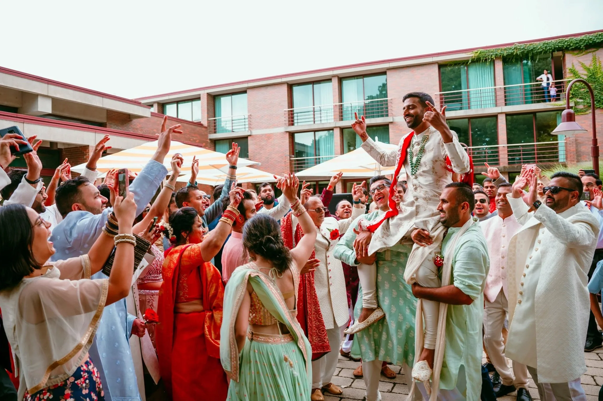 People celebrating at an outdoor wedding, with a groom being lifted on a man's shoulders, surrounded by guests in colorful traditional attire.