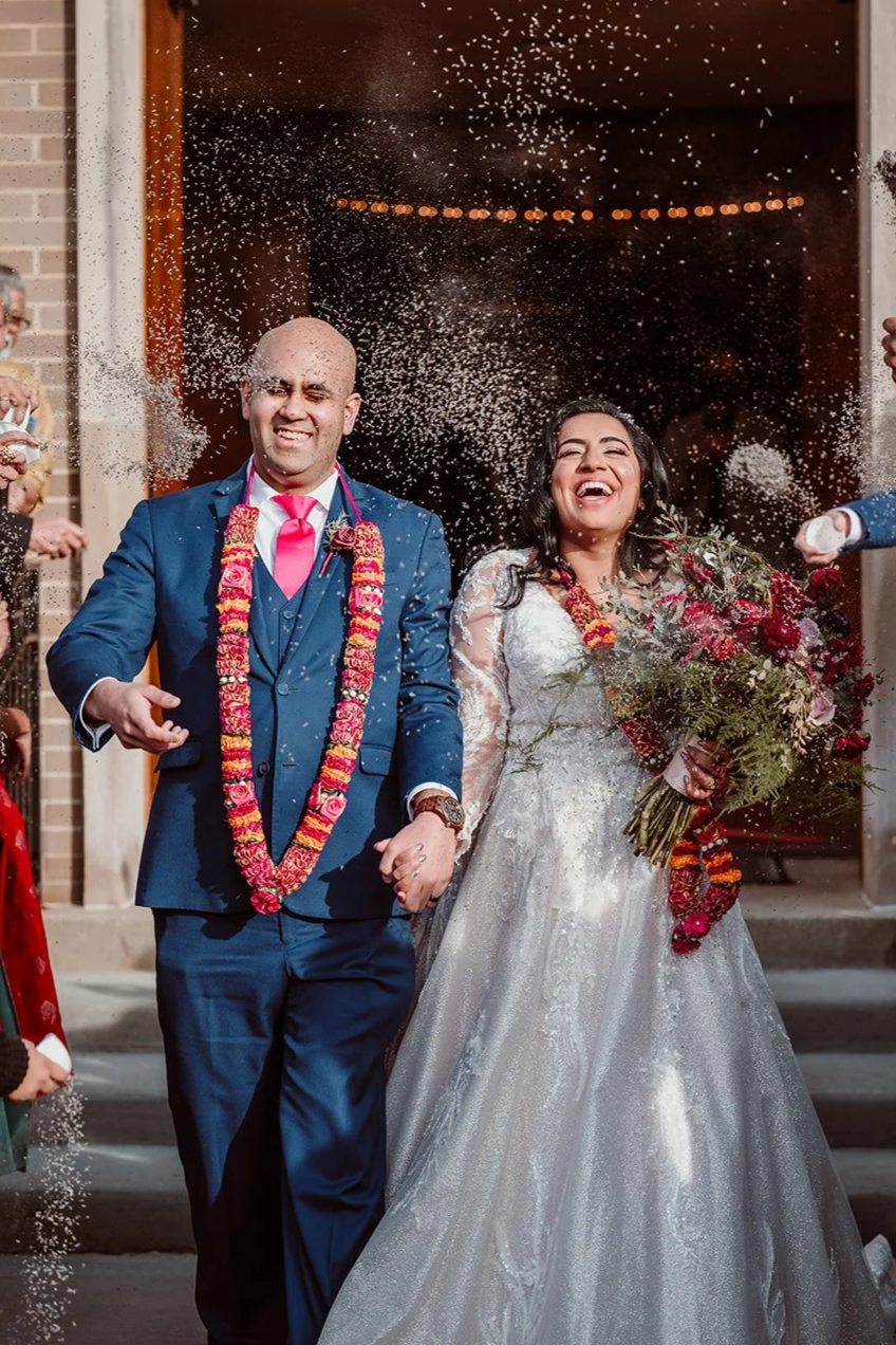 A newlywed couple celebrating outside a building, spraying rice or confetti. The groom is in a blue suit with a pink tie and a floral garland, and the bride is wearing a white wedding gown and holding a bouquet of flowers, both smiling joyfully.