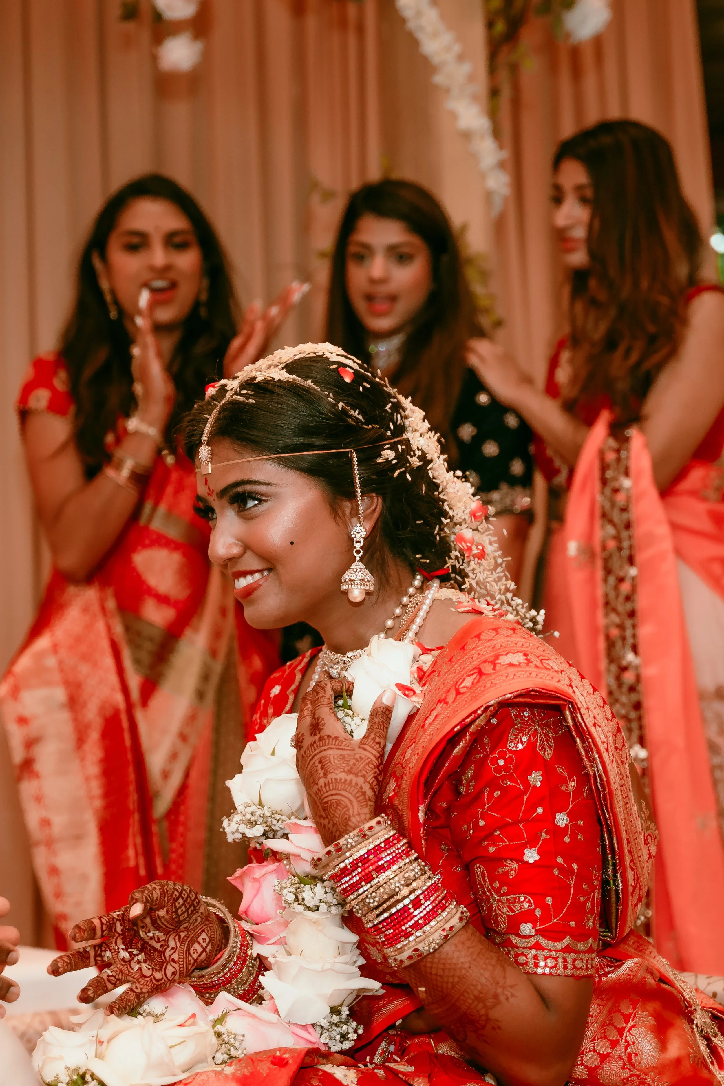 A bride in traditional Indian attire with henna on her hands, wearing jewelry and a red sari, holding a bouquet of white roses, surrounded by friends during her wedding.