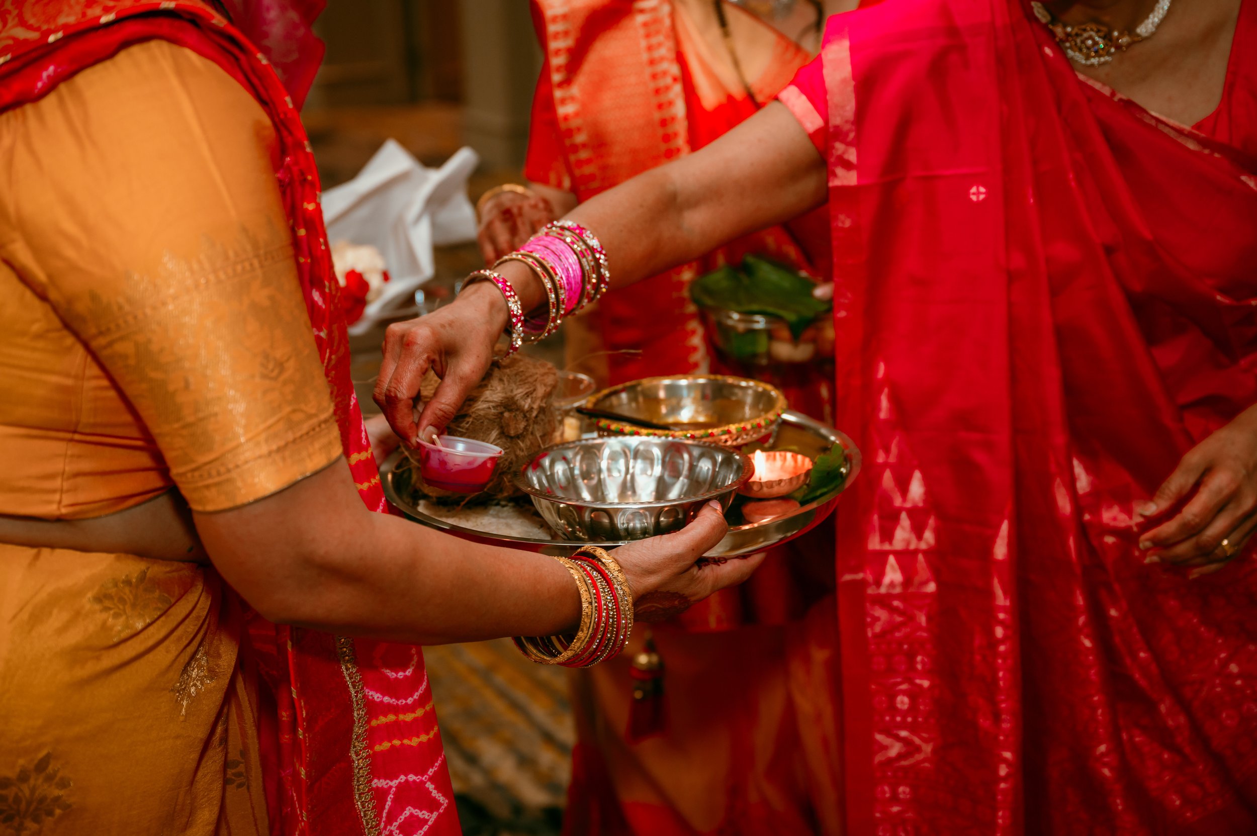 Women dressed in traditional Indian attire performing a religious ritual, offering a red liquid in small cups on a silver tray.