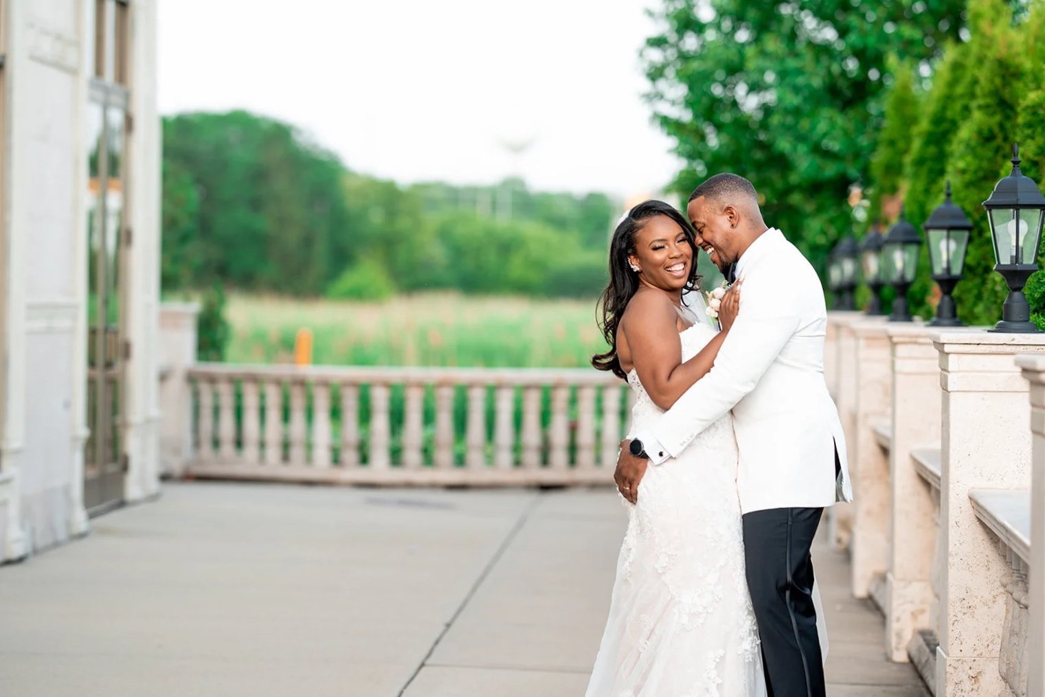 A happy couple on their wedding day, embracing each other and smiling, outdoors on a balcony with greenery in the background.