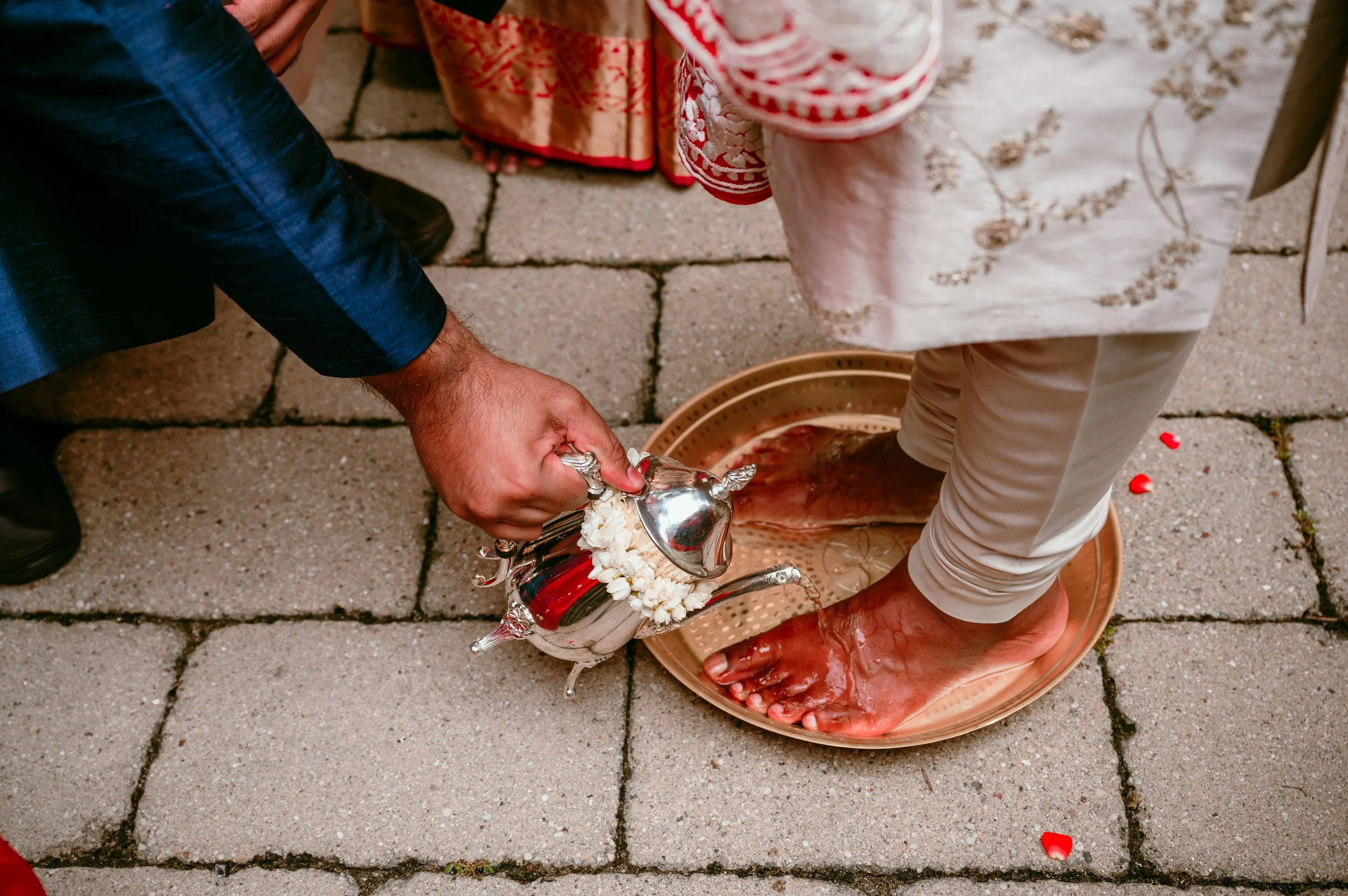 Foot washing ceremony with person washing another person's feet with water from a silver pitcher, on a tiled ground.