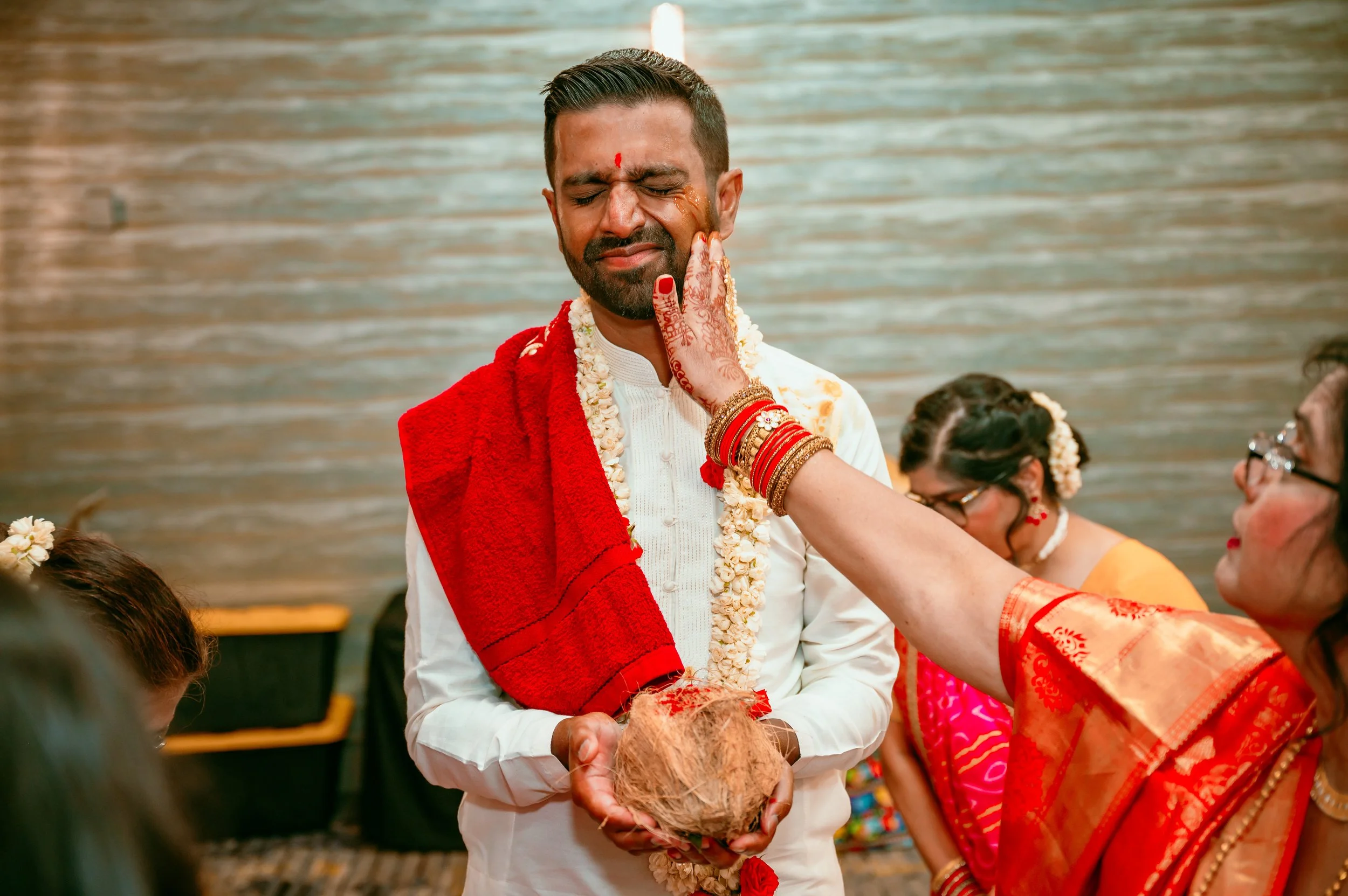 Man with closed eyes receiving an Aarti ceremony during a traditional Indian wedding, holding a coconut in his hands, wearing a white kurta with garlands, while a woman applies turmeric on his face.