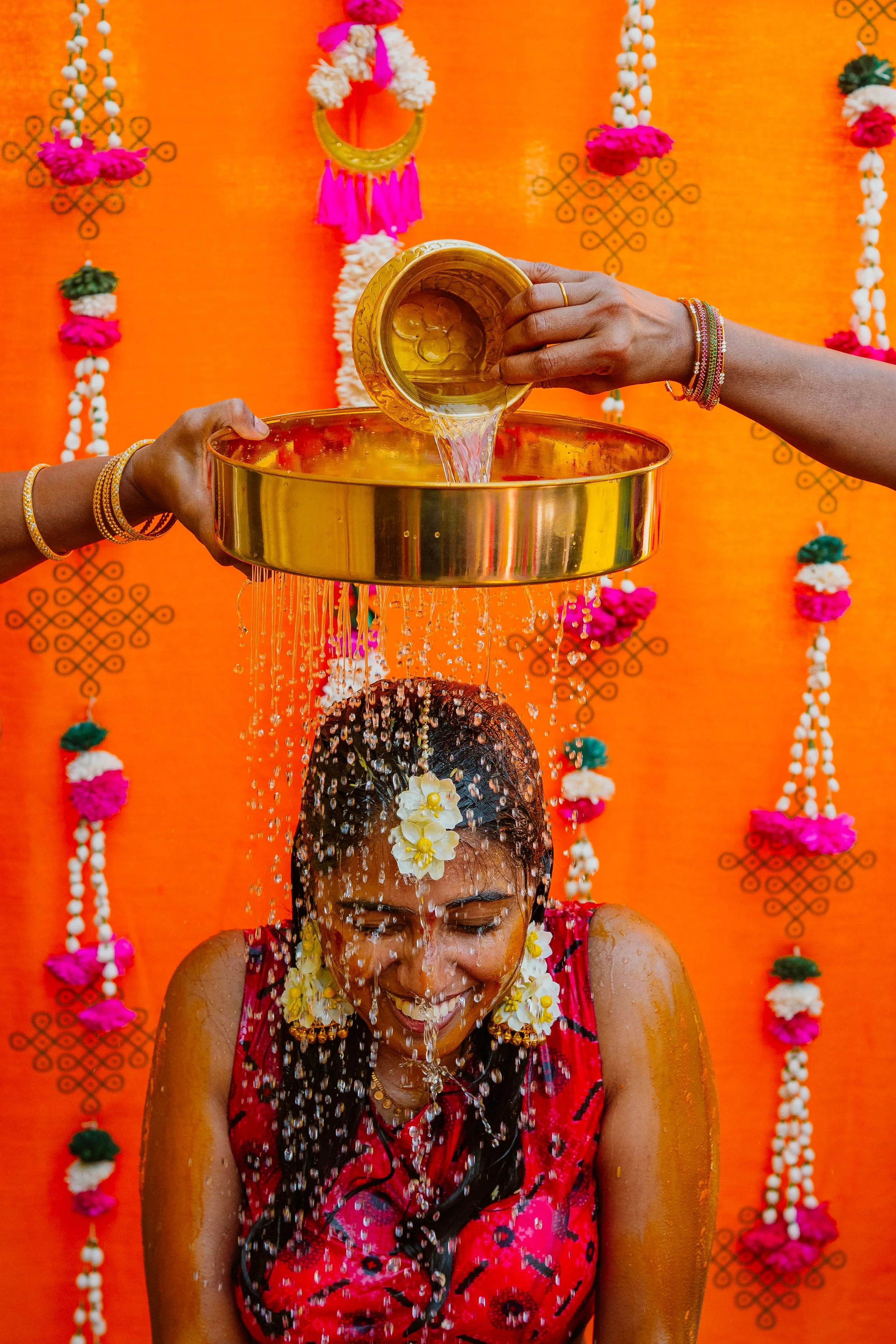A woman participating in a traditional Indian ritual, pouring water or liquid over her head in front of a vibrant orange background decorated with floral ornaments.