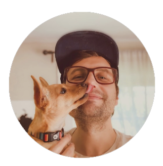 Man with glasses and a cap being licked by a small dog.