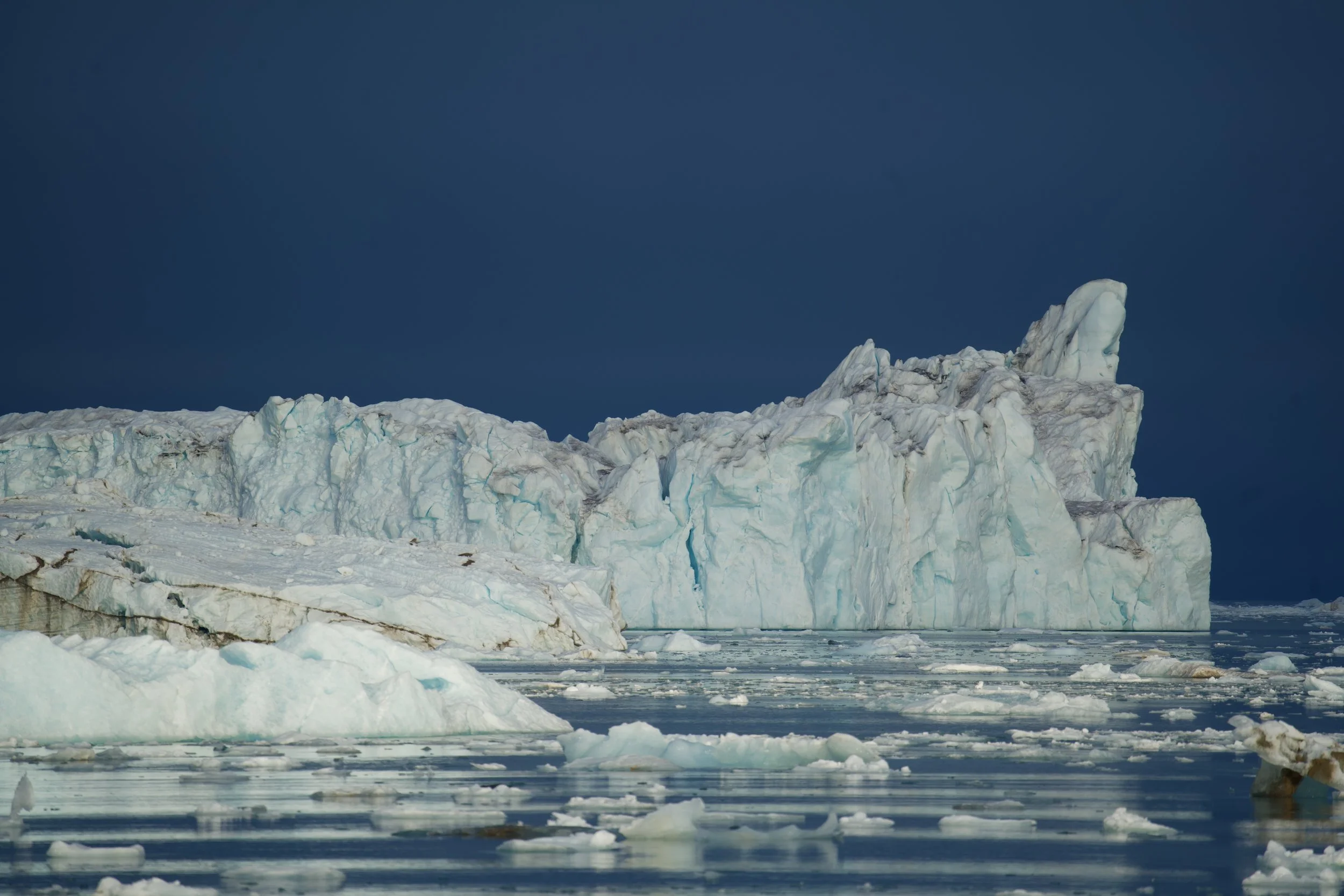 Sunset over a large glacier calving into the ocean with ice falling into the water and waves splashing.
