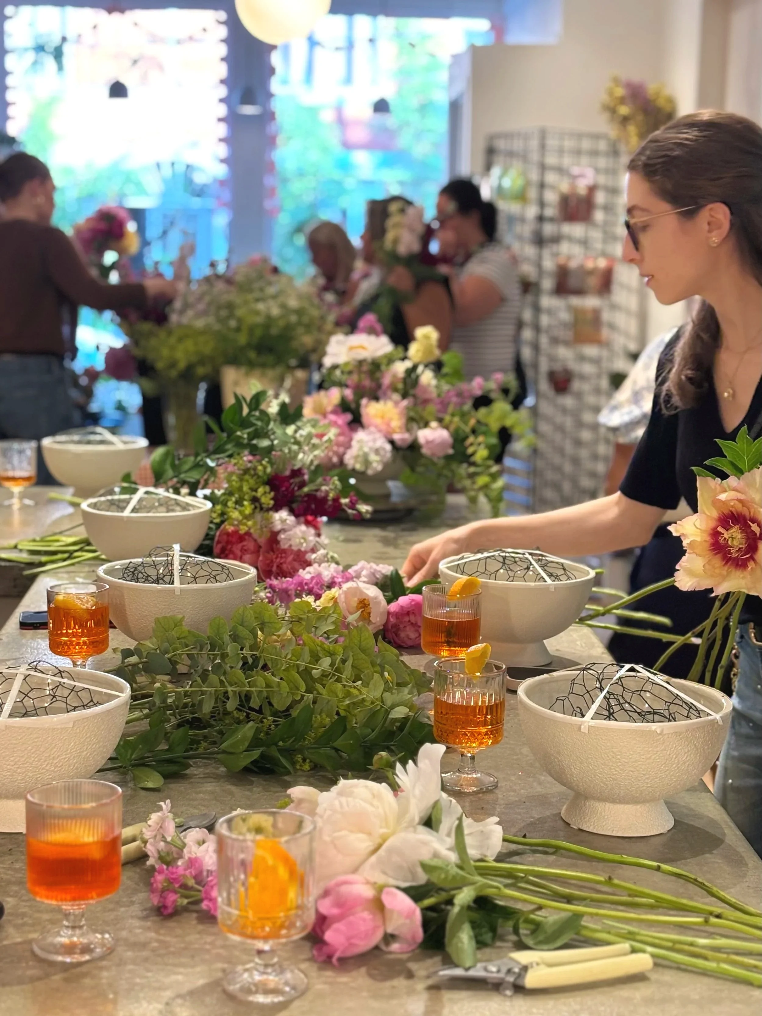 A group of women gathered around a table arranging flowers, with various floral arrangements, flowers, and glasses of orange-colored drinks. The setting appears to be indoors, possibly a floral workshop or class.
