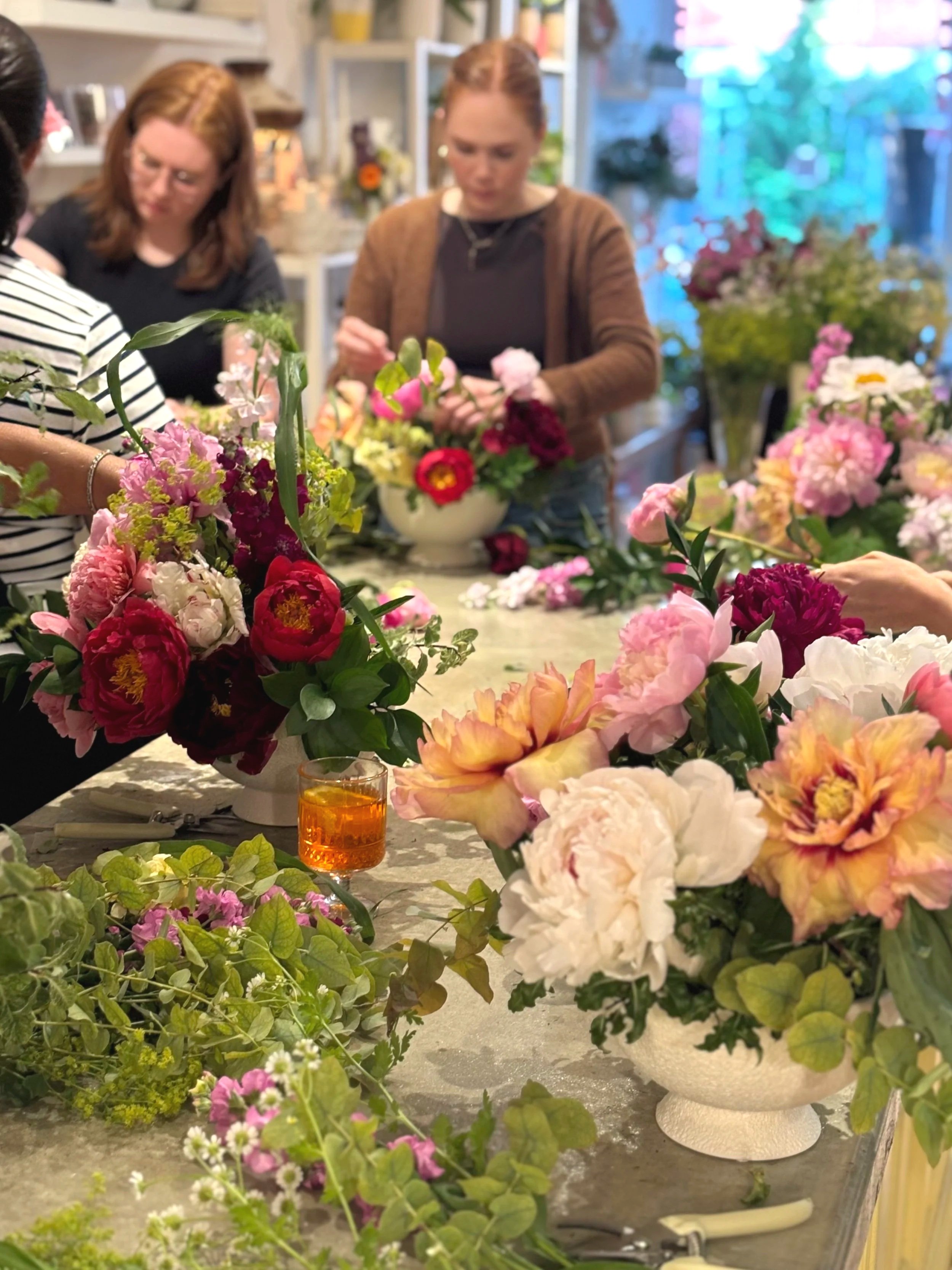 People gathered around a table with various colorful flowers in vases, preparing floral arrangements in a bright room.