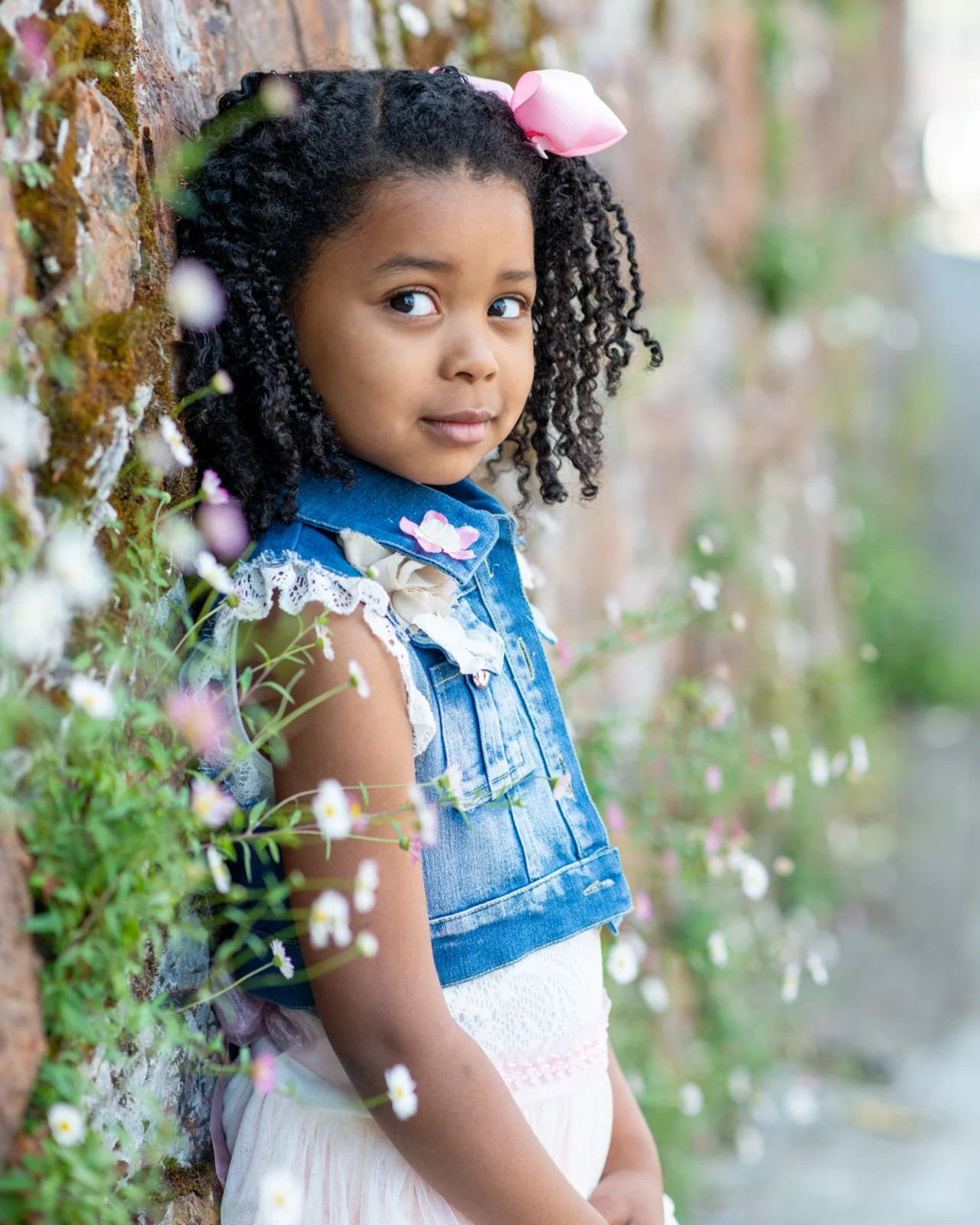 A young girl with curly hair wearing a pink bow and denim vest standing next to a mossy brick wall surrounded by small flowers.