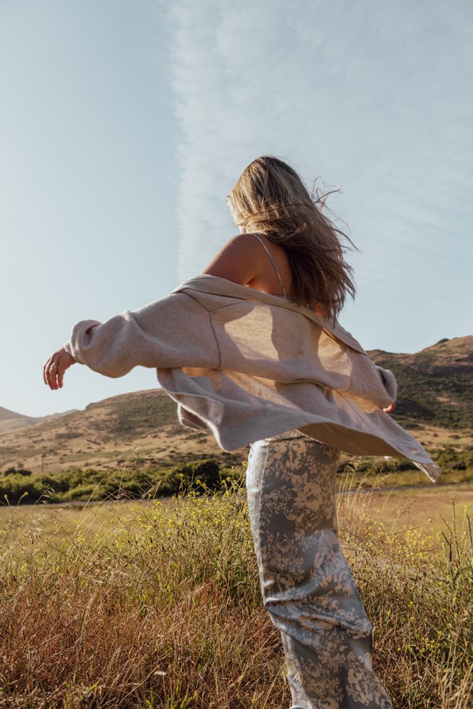 Woman with long hair standing in an open field with hills in the background, wearing a beige jacket and patterned pants, with the sky overhead.