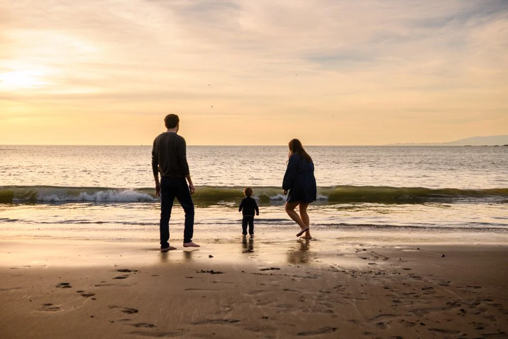 A family of three on a beach at sunset, with a man, a woman, and a small child near the water's edge.