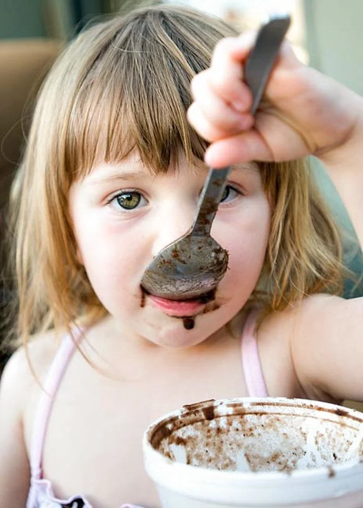 Young girl with light brown hair and green eyes eating chocolate ice cream with a spoon, holding it above her mouth, with a bowl of ice cream in front of her