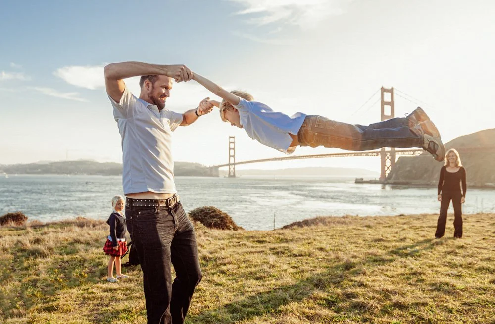 A man and a boy are playing with a toy in front of the Golden Gate Bridge, with two women and water in the background.