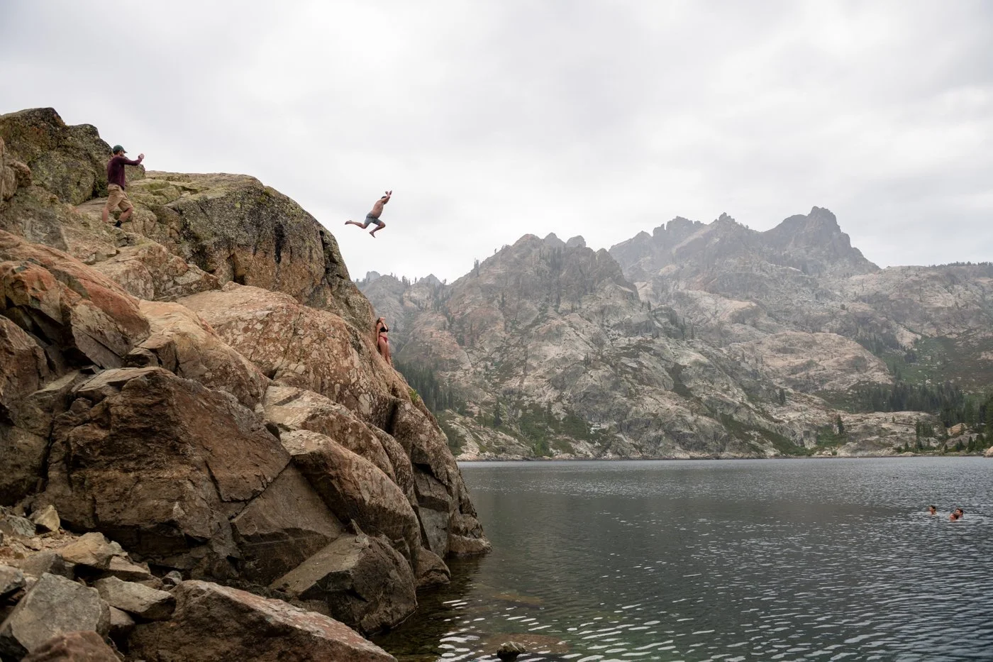 People jumping off rocks into a lake surrounded by mountainous terrain under a cloudy sky.