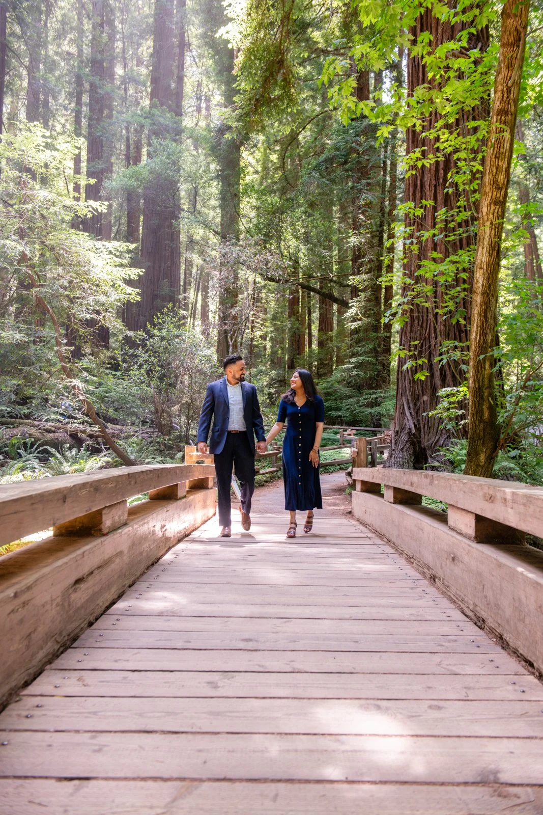 A couple dressed in formal attire walking hand in hand on a wooden bridge in a lush forest with tall trees and green foliage.