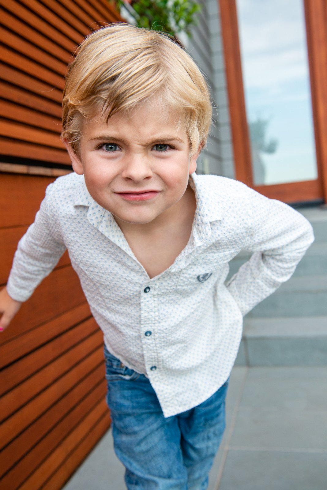 Close-up of a young boy with blonde hair and blue eyes, wearing a white button-up shirt with a small pattern and blue jeans, leaning forward outdoors in front of a wooden wall and a glass door.