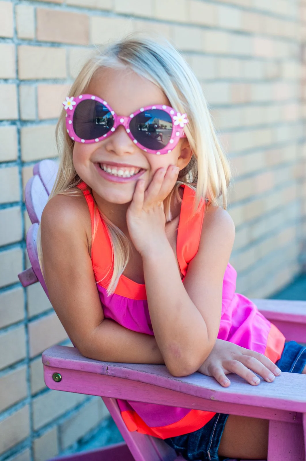 A young girl smiling and wearing pink sunglasses with flower decorations, sitting on a pink bench next to a brick wall, dressed in a bright pink and orange sleeveless top.