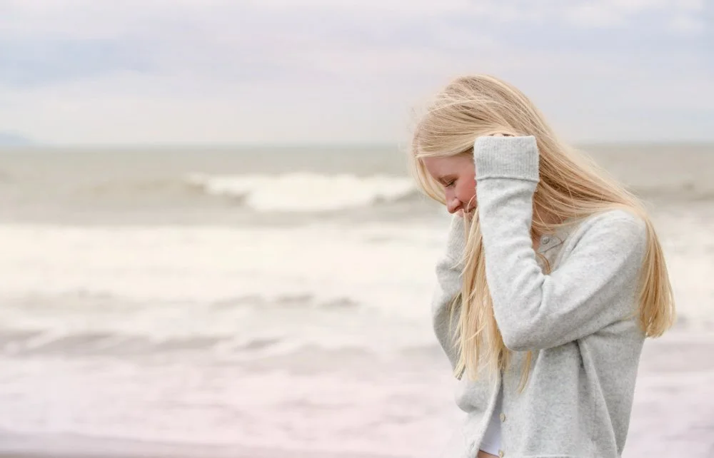 A blonde girl in a gray hoodie standing on a beach, looking downward with a smile.