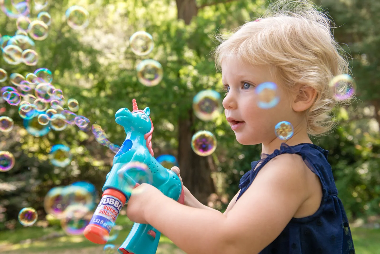 Young blonde girl in a navy dress blowing bubbles outdoors with a blue unicorn bubble blower.