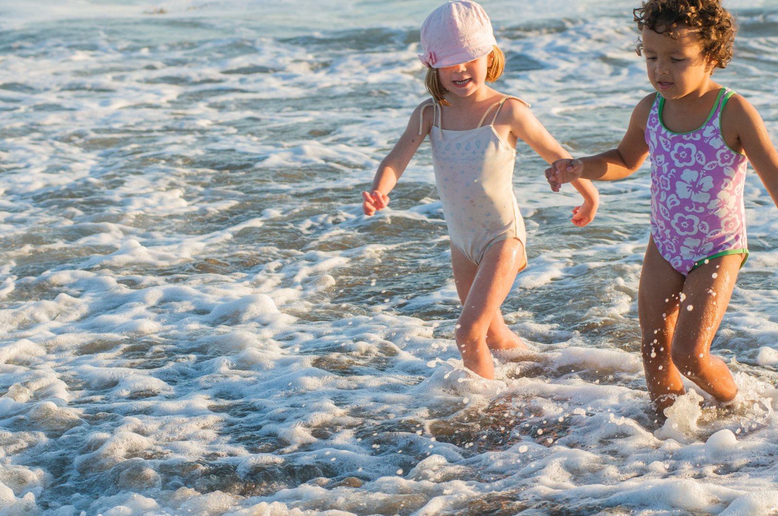 Two young girls playing in the surf at the beach, wearing swimsuits and holding hands.