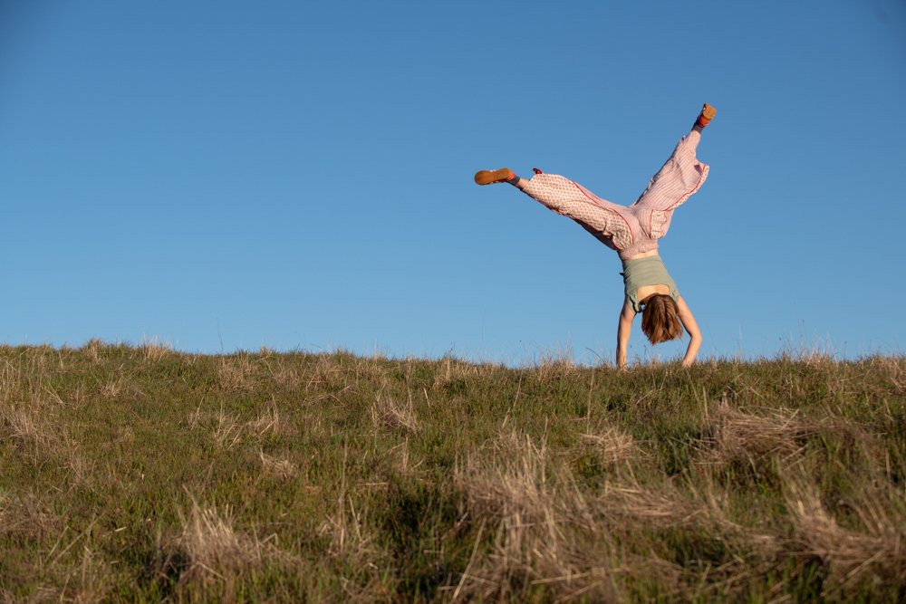 A person performing a handstand on a grassy hill under a clear blue sky.