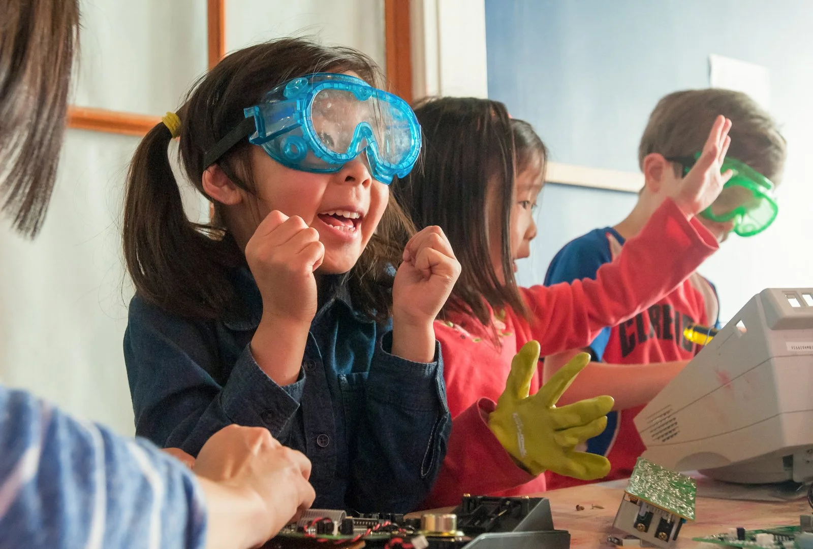 Children wearing protective goggles and gloves working with electronic components and a device at a science activity or classroom.