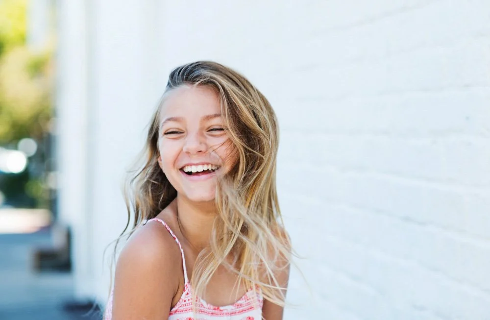A young girl with long blonde hair smiling and laughing outdoors near a white brick wall.