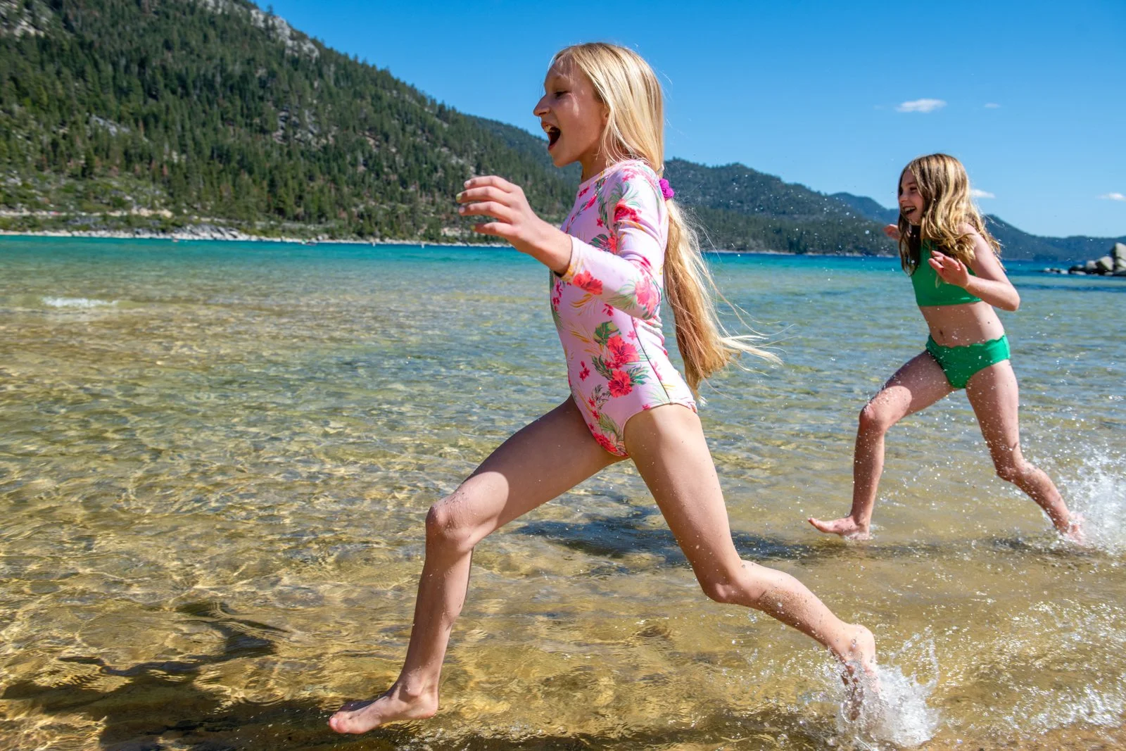 Two young girls running and playing in shallow water at a beach with mountains in the background.