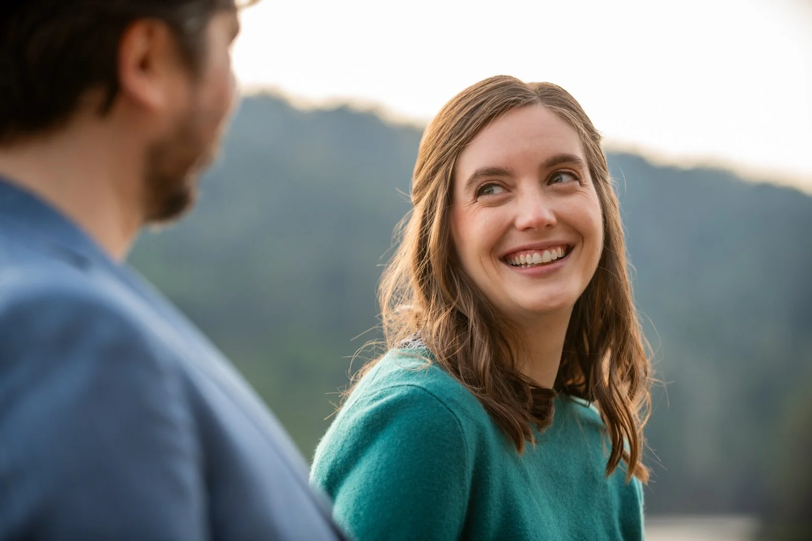 A woman with brown hair smiling and looking at a man who is partially visible, against a blurred outdoor background.