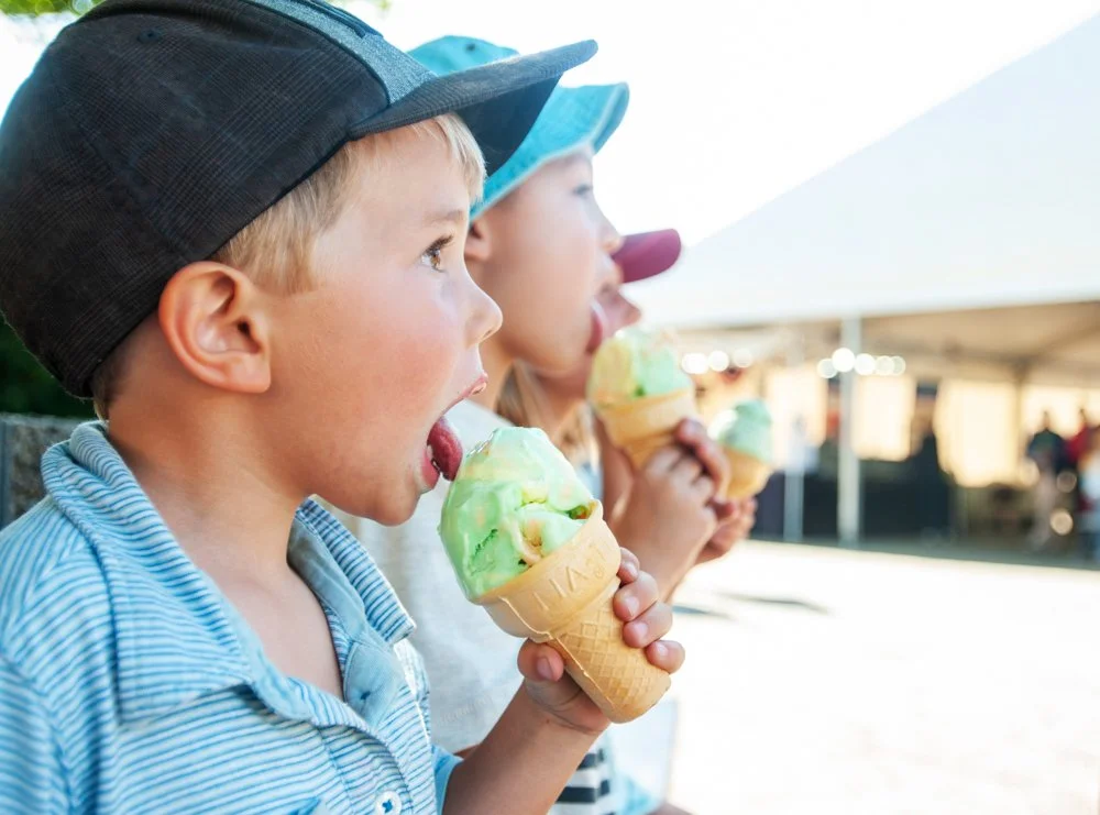 Three children wearing caps eating ice cream cones outside under a tent.
