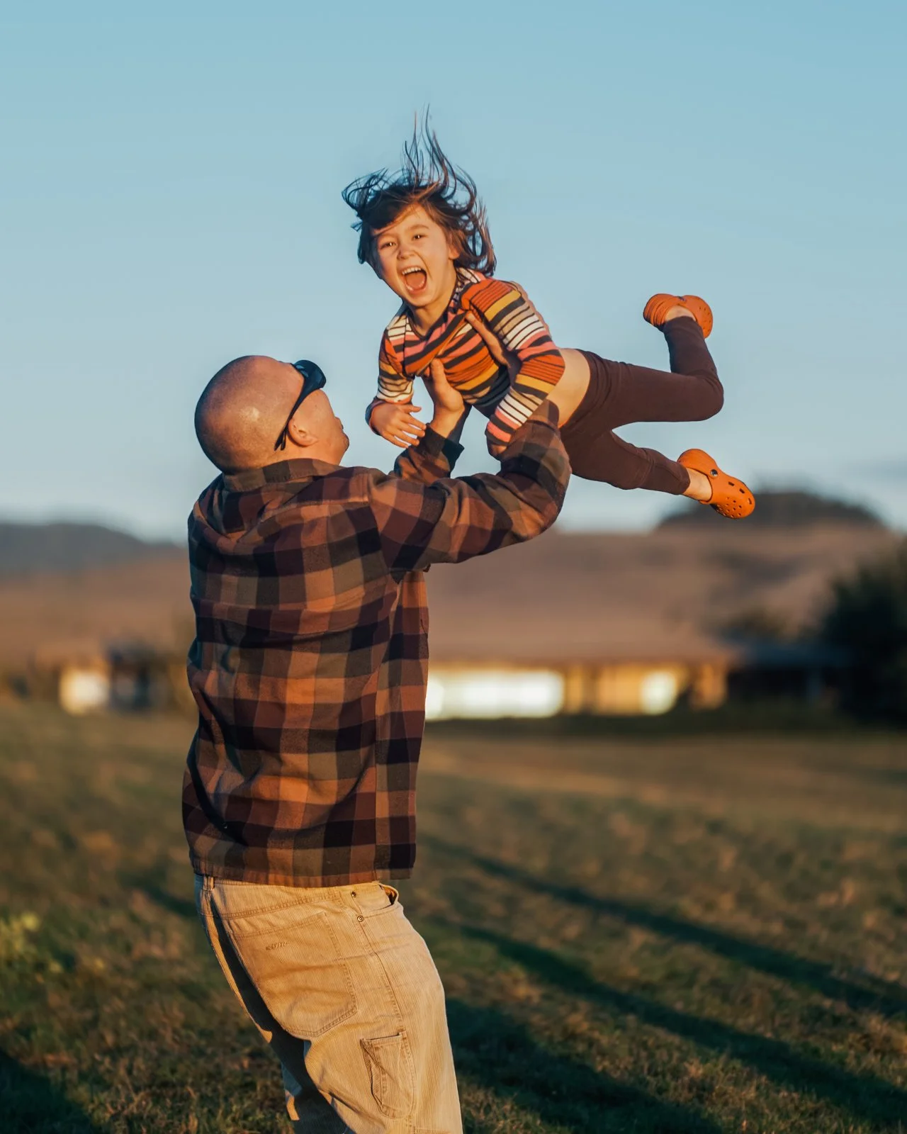 A man in a plaid shirt and khaki pants lifts a young girl in a striped shirt and brown pants into the air outdoors during sunset, with a background of open fields and a clear sky.