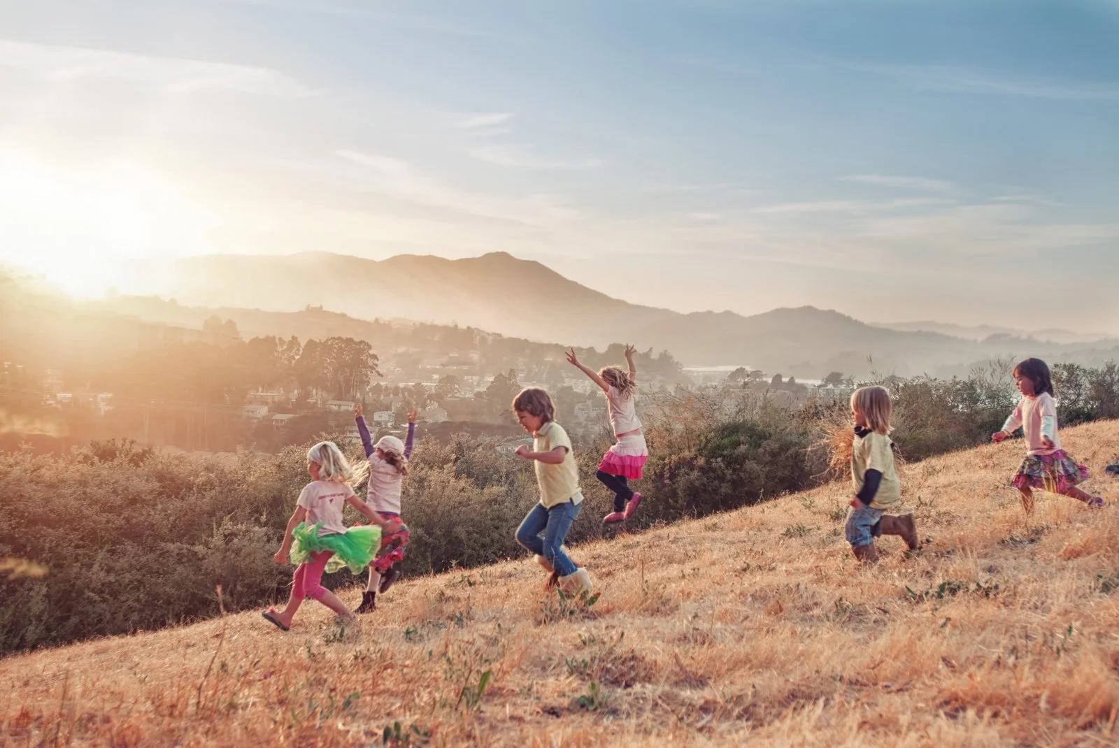 Children playing and jumping on a grassy hillside during sunset with mountains in the background.