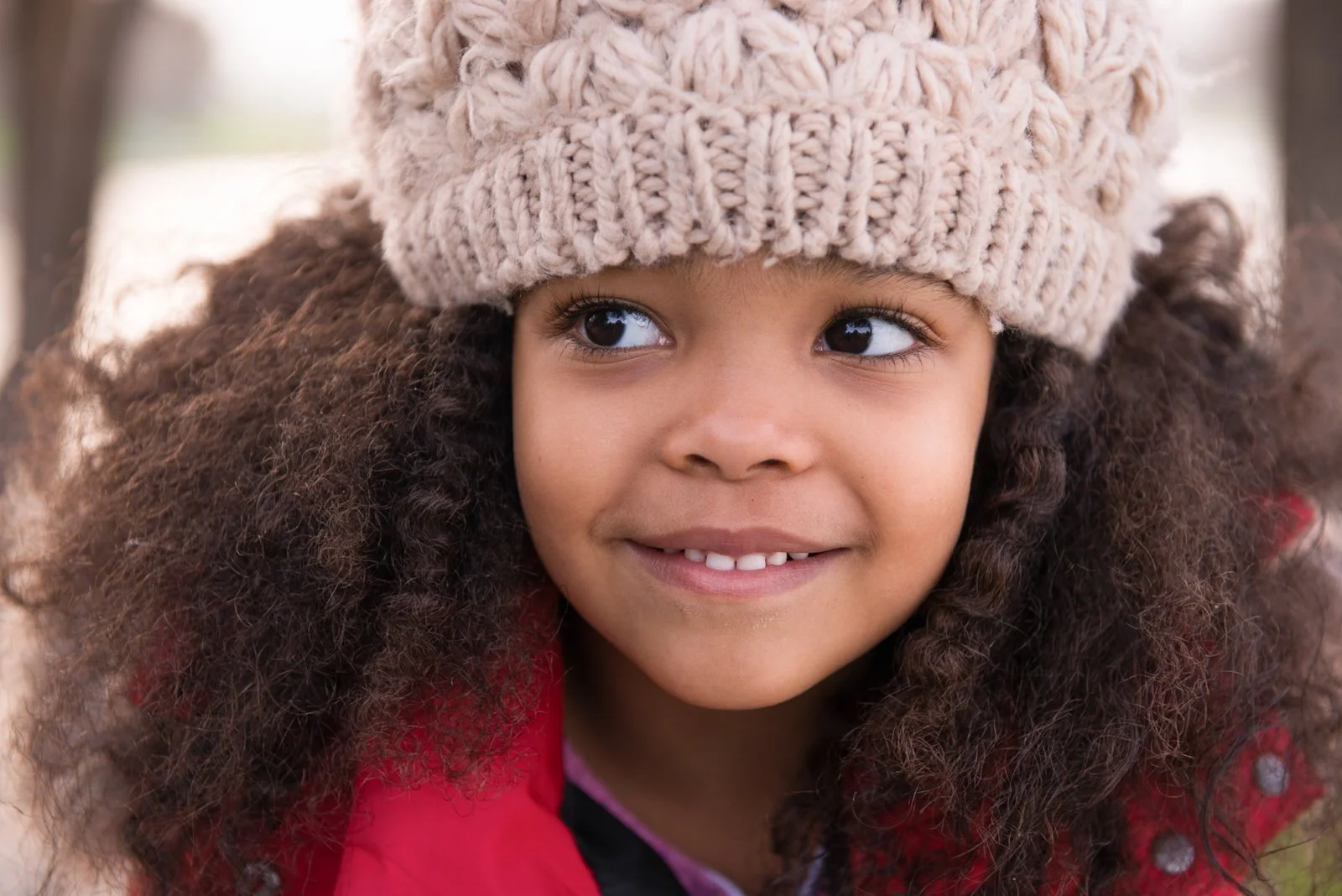 Close-up portrait of a young girl with curly hair wearing a beige knit hat and red jacket, smiling outdoors.