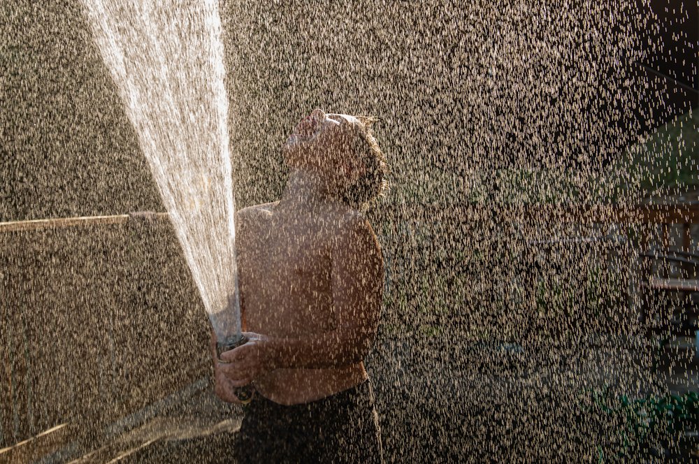 A person is standing outdoors in daylight while being sprayed with water from a garden hose.