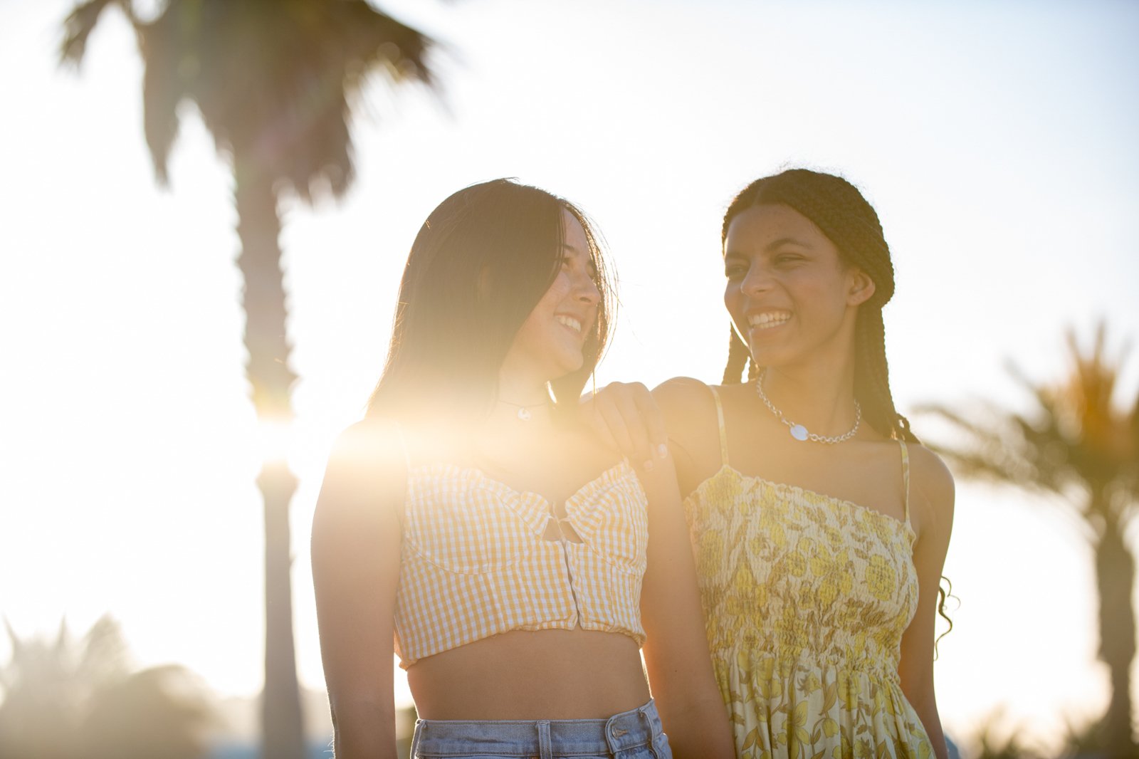 Two young women with dark hair sharing a joyful moment outdoors with palm trees in the background, backlit by the sun, one wearing a yellow checkered crop top and the other in a floral yellow dress.